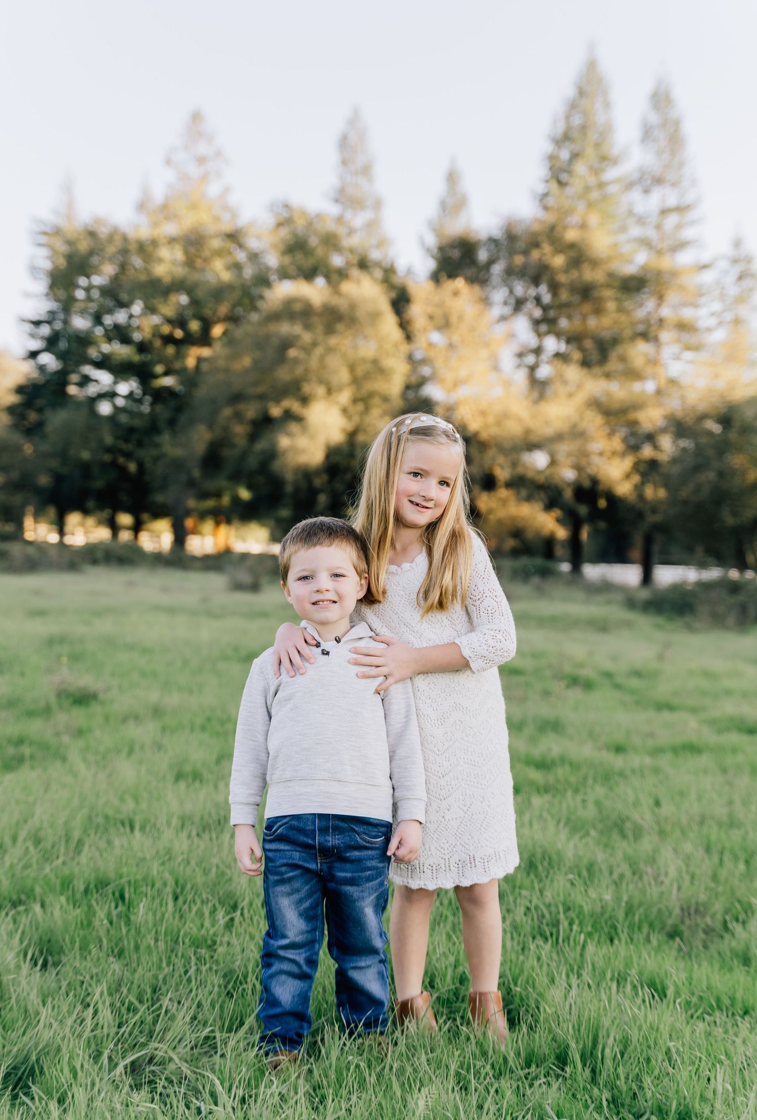 Two children play together in a grassy field on a sunny day with trees in the background.