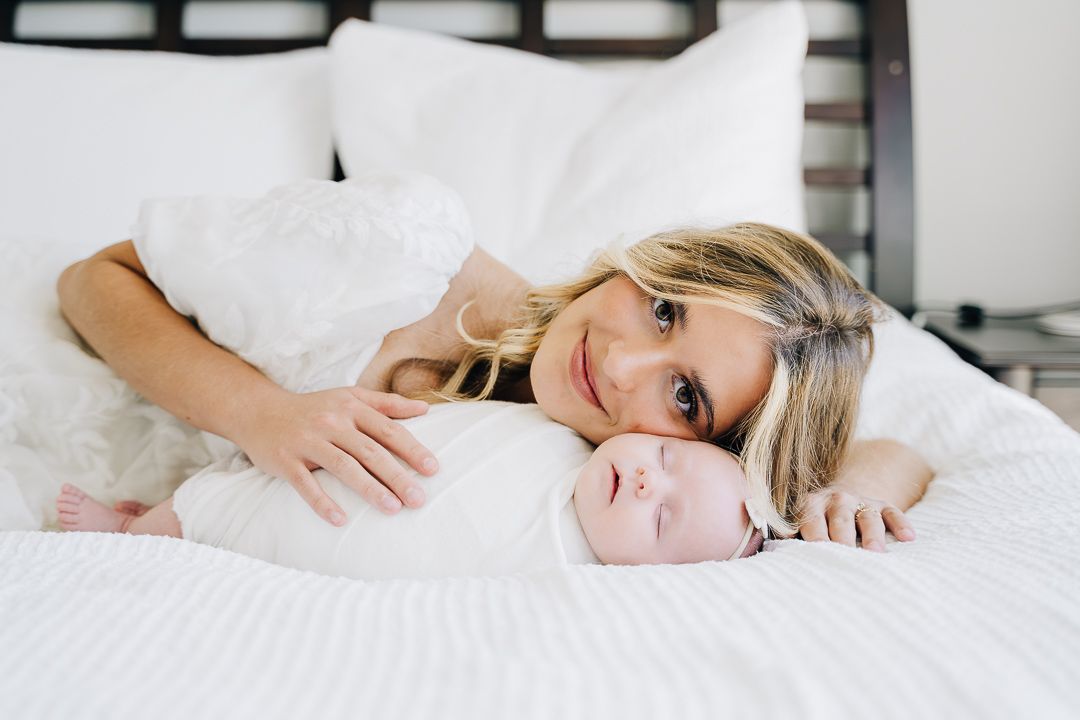 Smiling blonde mother lying on white bed with her newborn baby in a tender bonding moment.