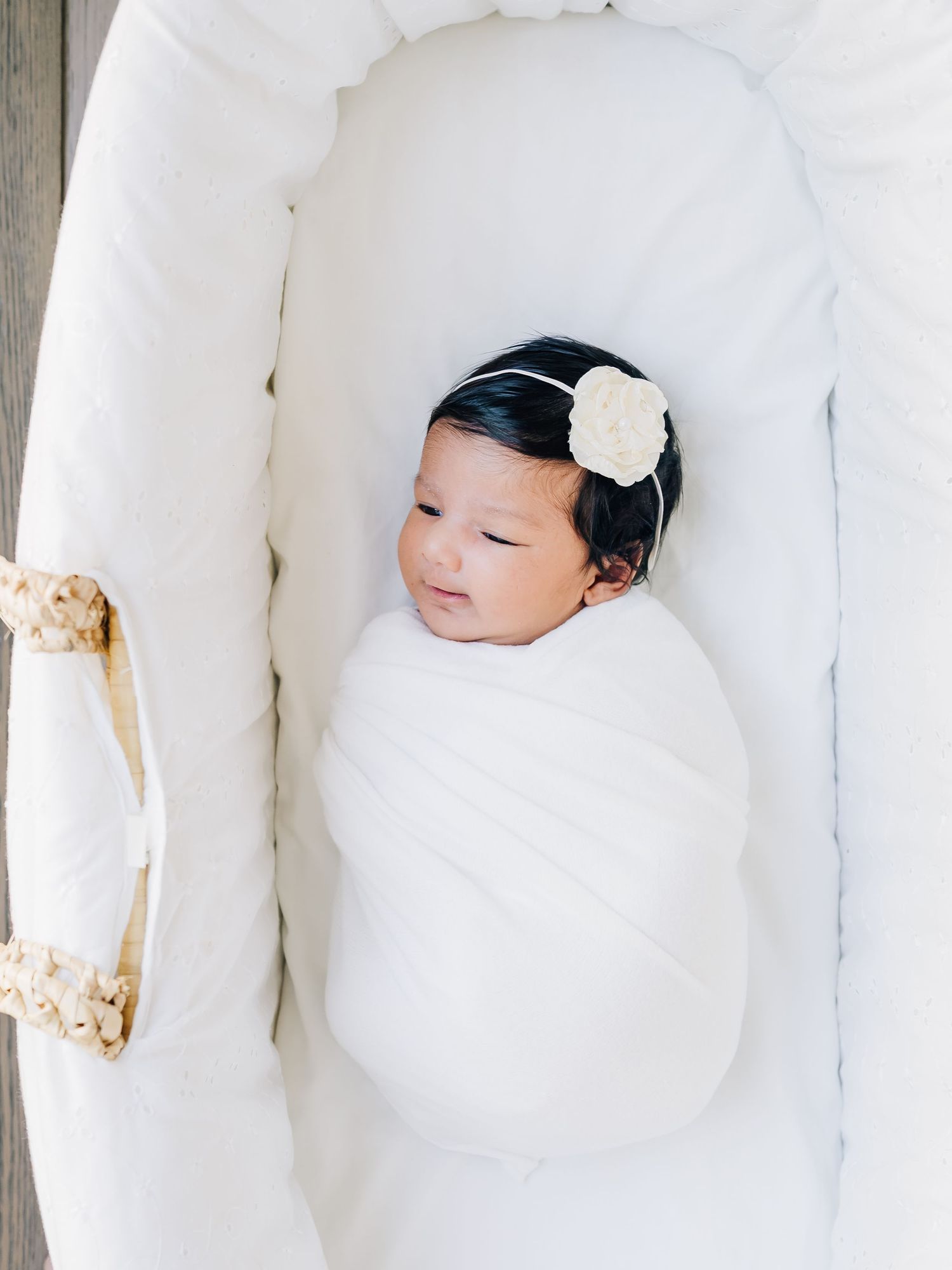 Newborn baby girl swaddled in white blanket with floral headband sleeping peacefully in a white bassinet.