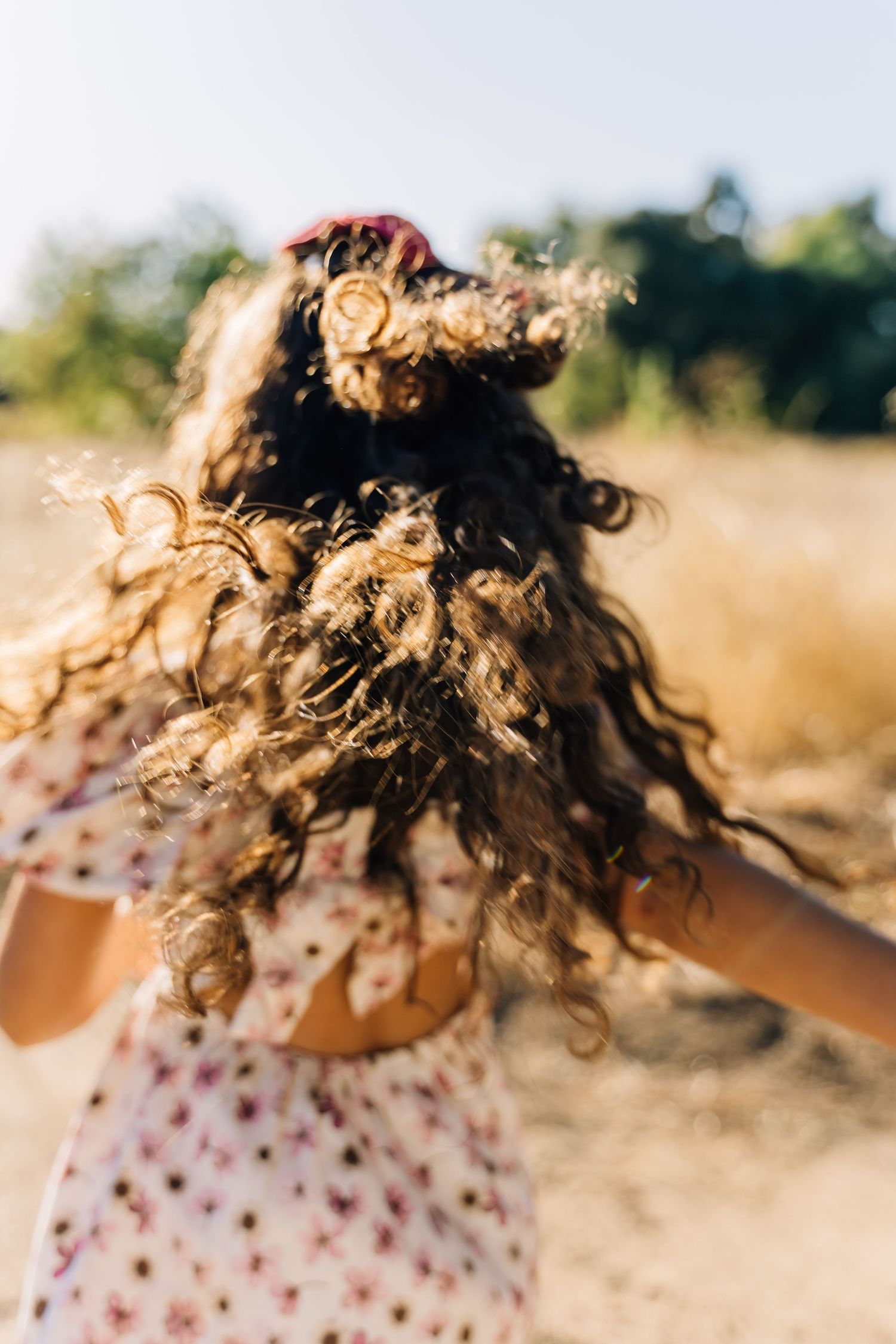 Curly brown hair blows in the wind against a sunny outdoor background with a floral patterned dress visible.