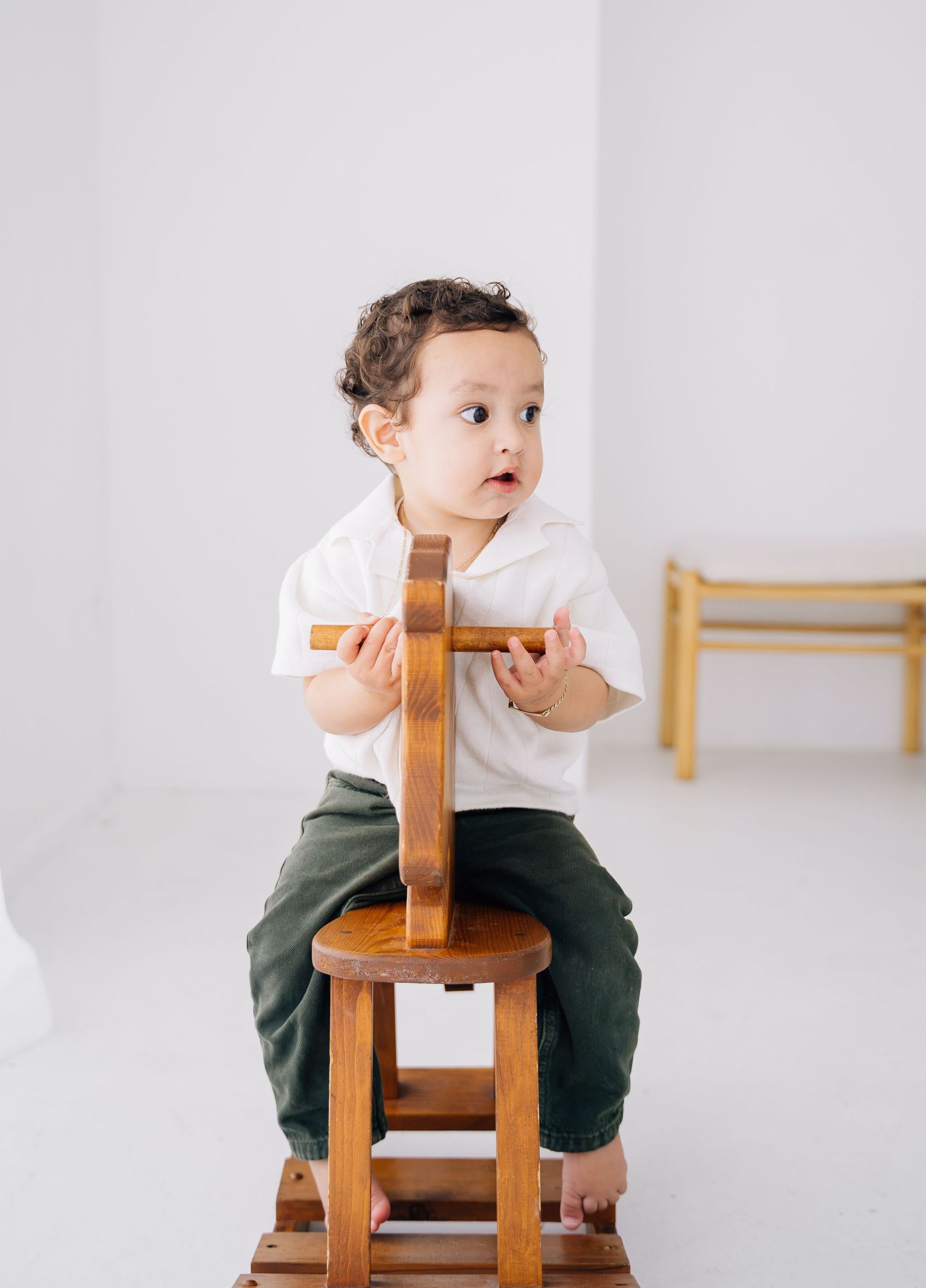 Cute toddler sitting on a wooden stool in a white studio, wearing a white shirt and green pants.