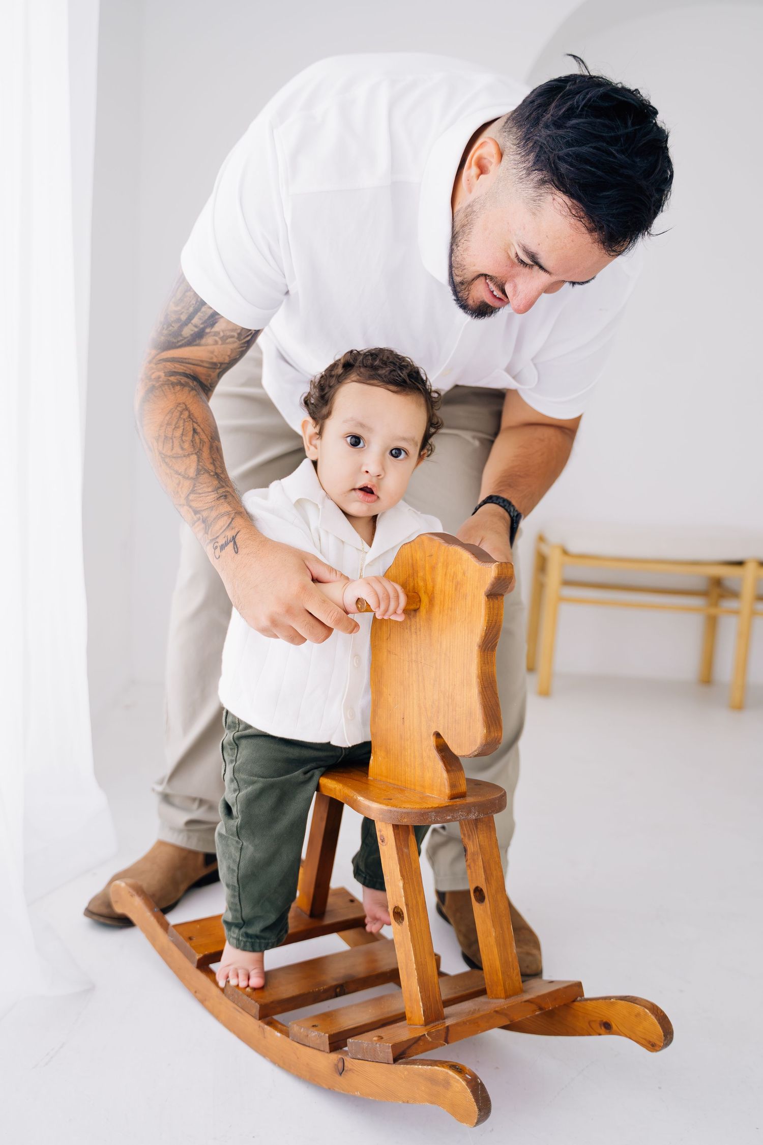 Tattooed father helping toddler ride a wooden rocking horse in a bright white room.