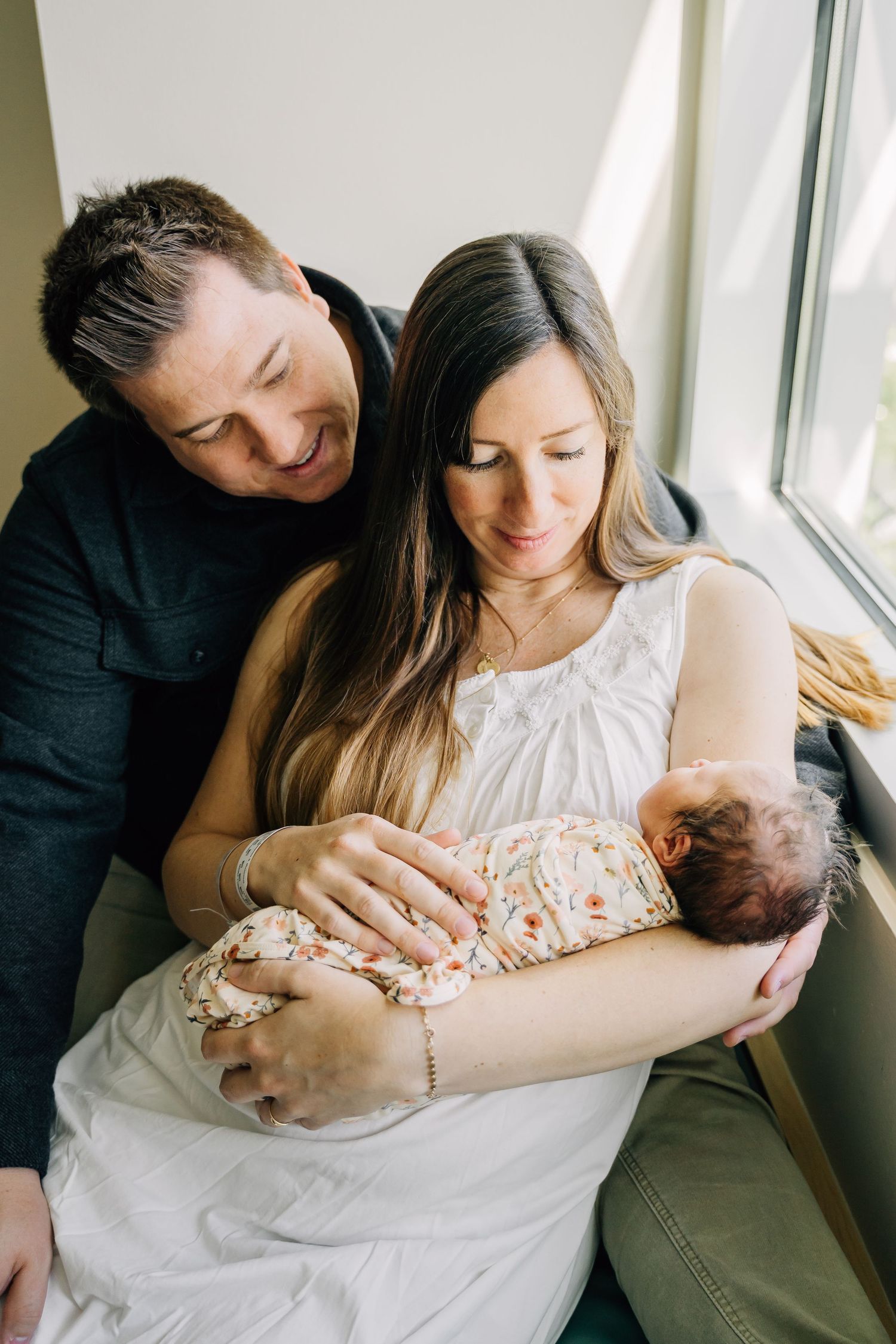 Happy couple smiling at their newborn baby wrapped in floral blanket near a bright window.