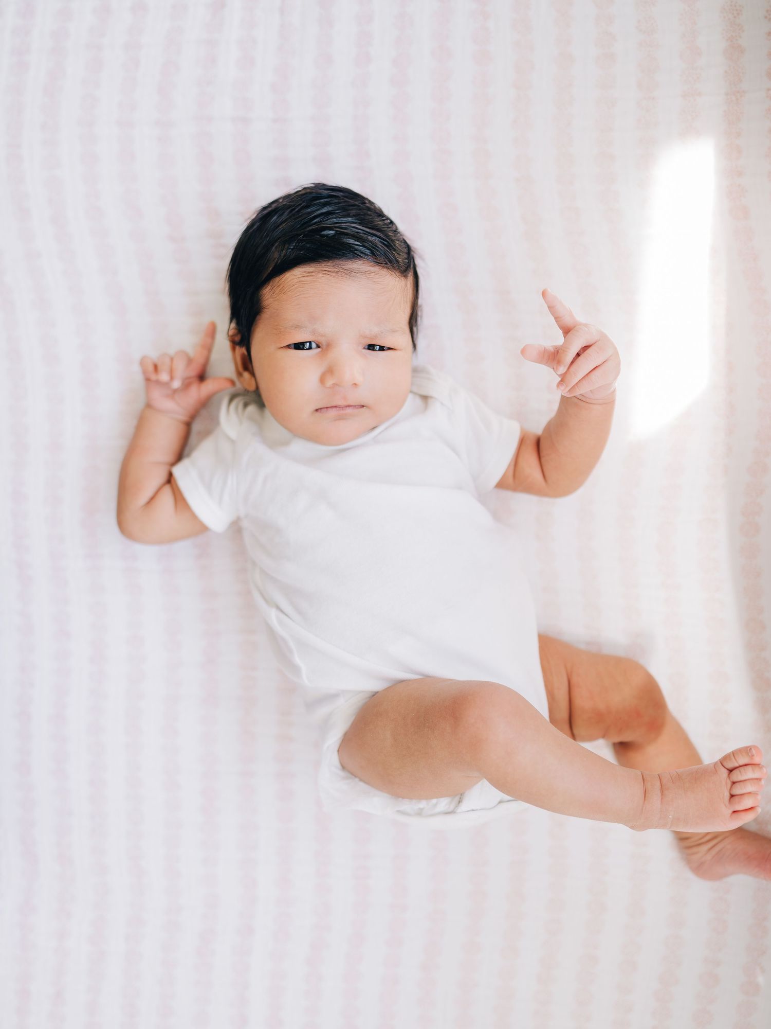 Adorable baby in white onesie lying on striped blanket with arms raised, looking at camera in soft natural light.