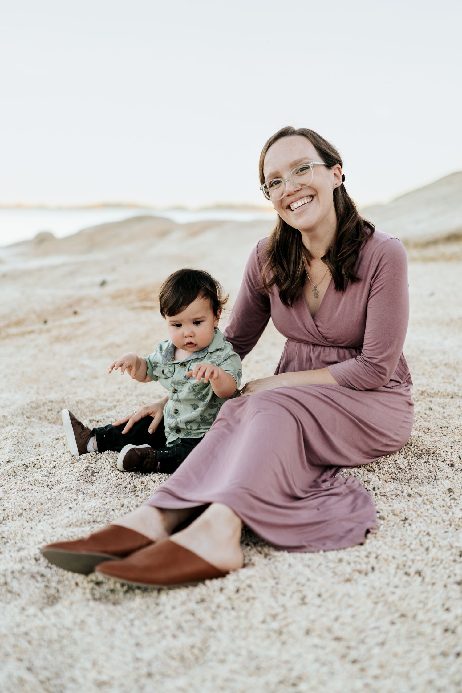 Family enjoying a relaxed moment together while sitting on sandy beach wearing mauve and green outfits.