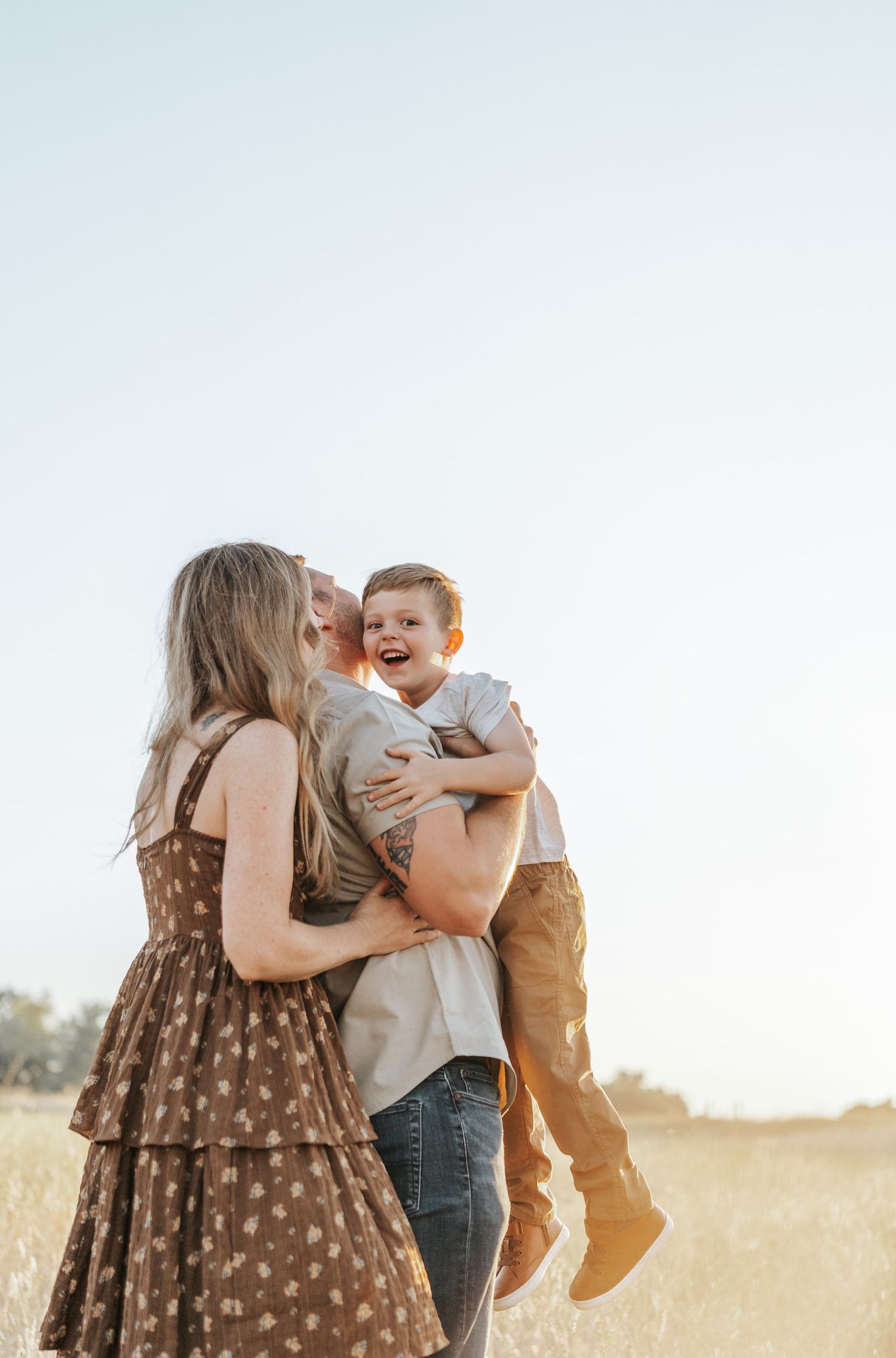 Family of three poses playfully in a sunlit meadow during golden hour, wearing casual outfits and brown polka dot dress.