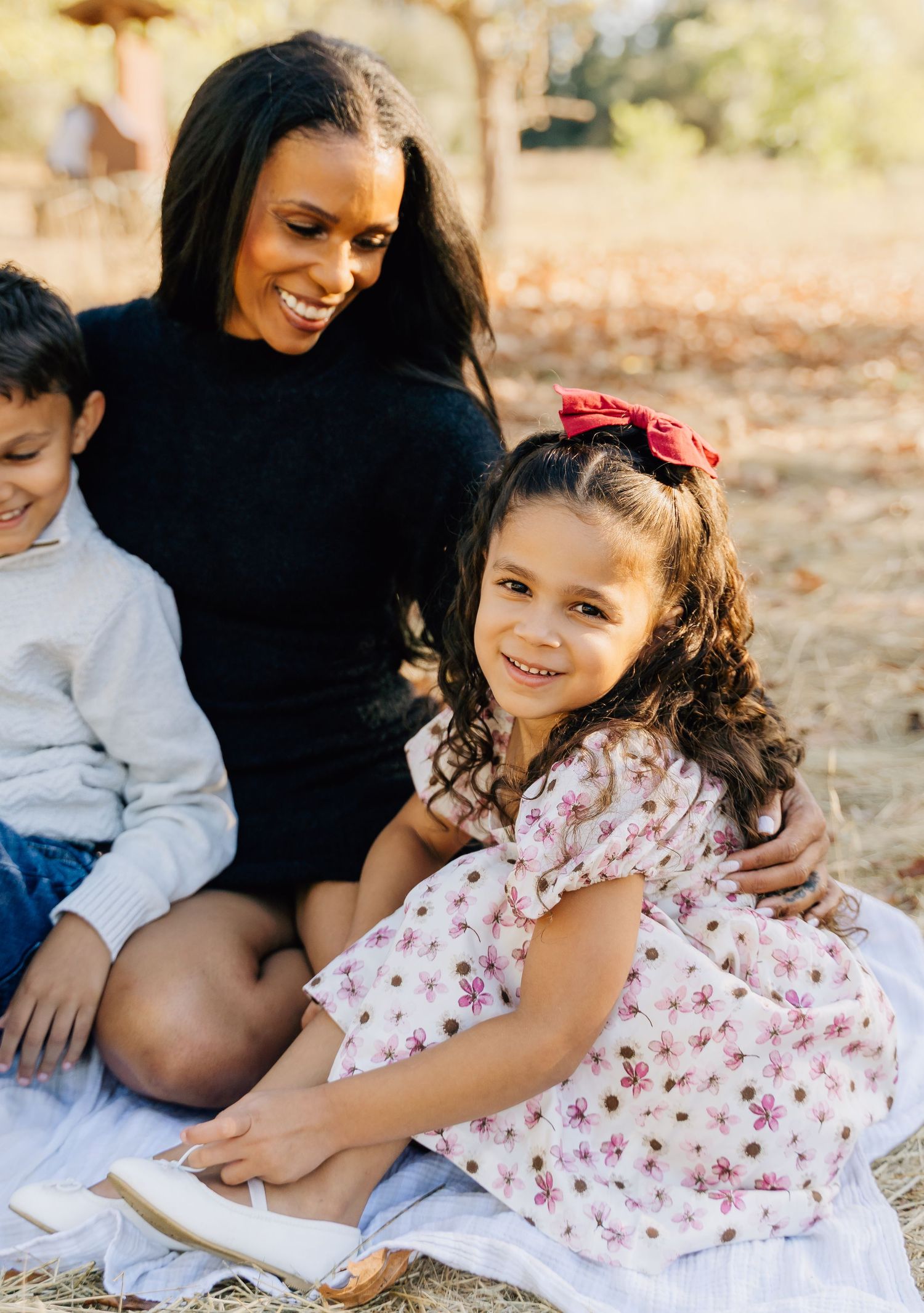 A joyful family moment outdoors with a woman in black and two children sitting on a blanket in autumn.