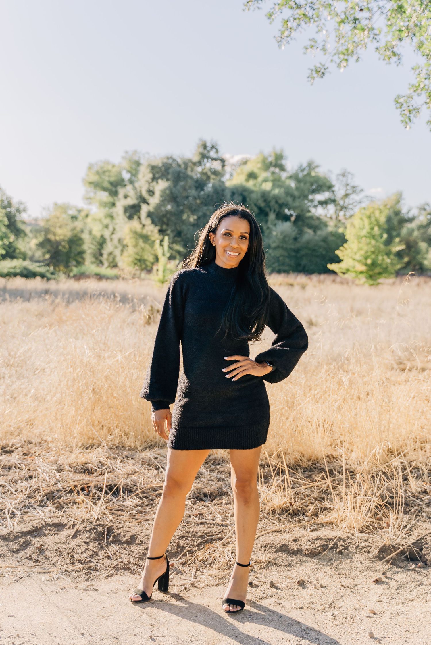 A person in a black sweater poses casually in a sunlit field with dried autumn grass.