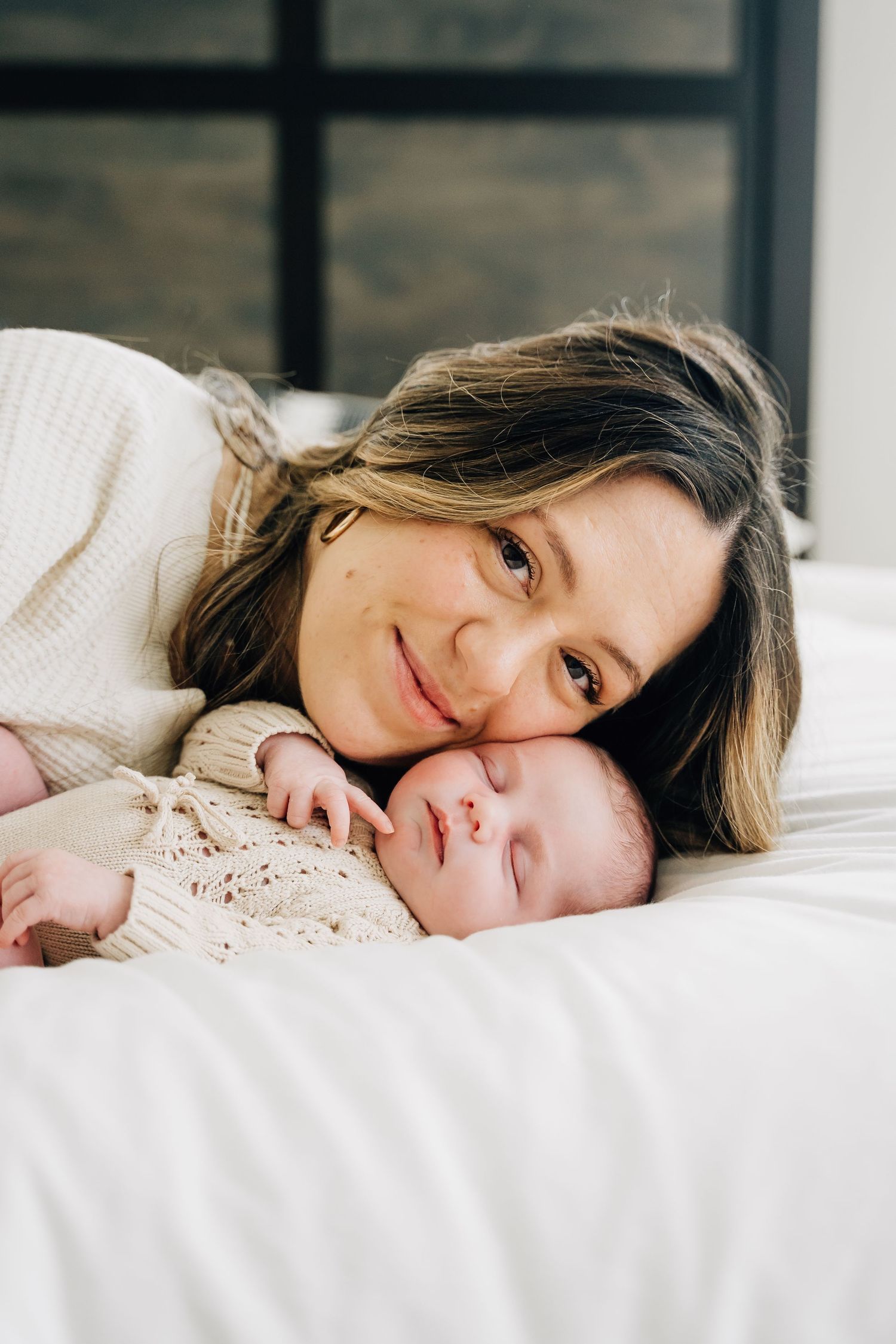 Smiling mother lying on white bed cheek-to-cheek with sleeping newborn baby dressed in cream knit outfit.
