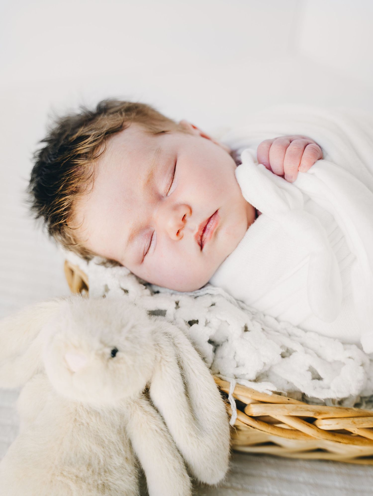 Peacefully sleeping newborn baby swaddled in white blanket resting in wicker basket with soft plush bunny toy.