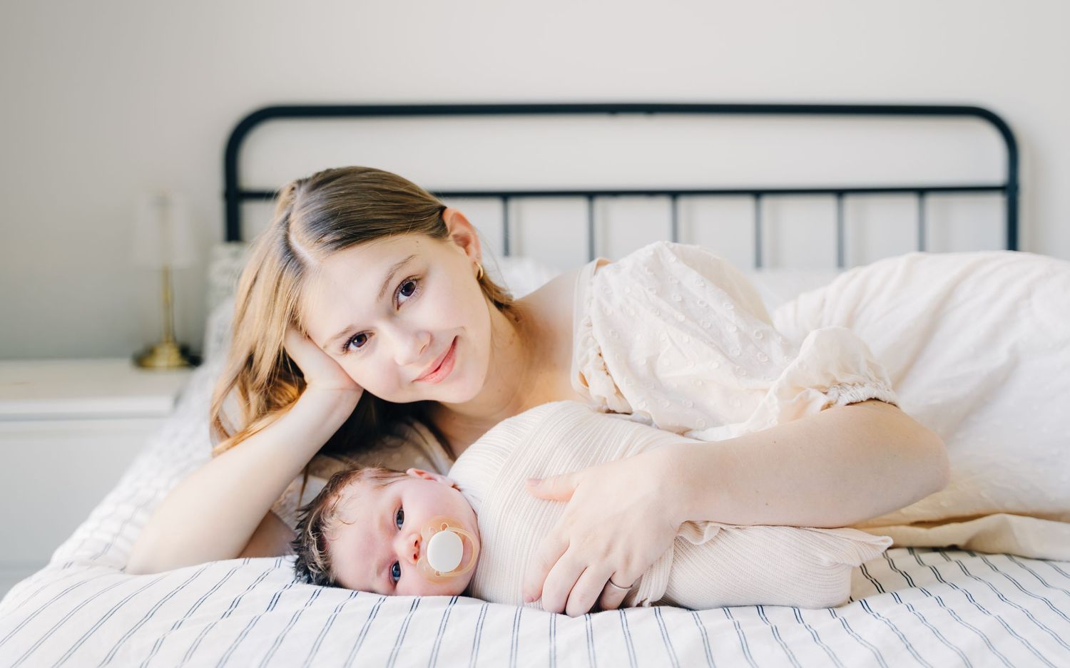 A smiling young mother lies on a white bed beside her sleeping newborn baby in a bright, cozy bedroom.
