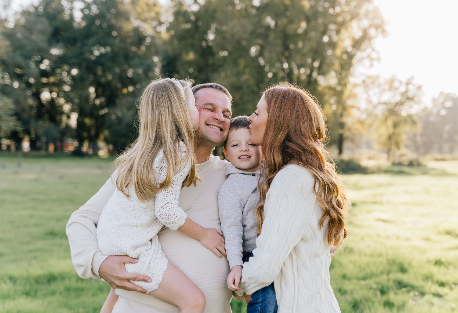 Family wearing coordinated white outfits embraces in a sunlit meadow during golden hour.