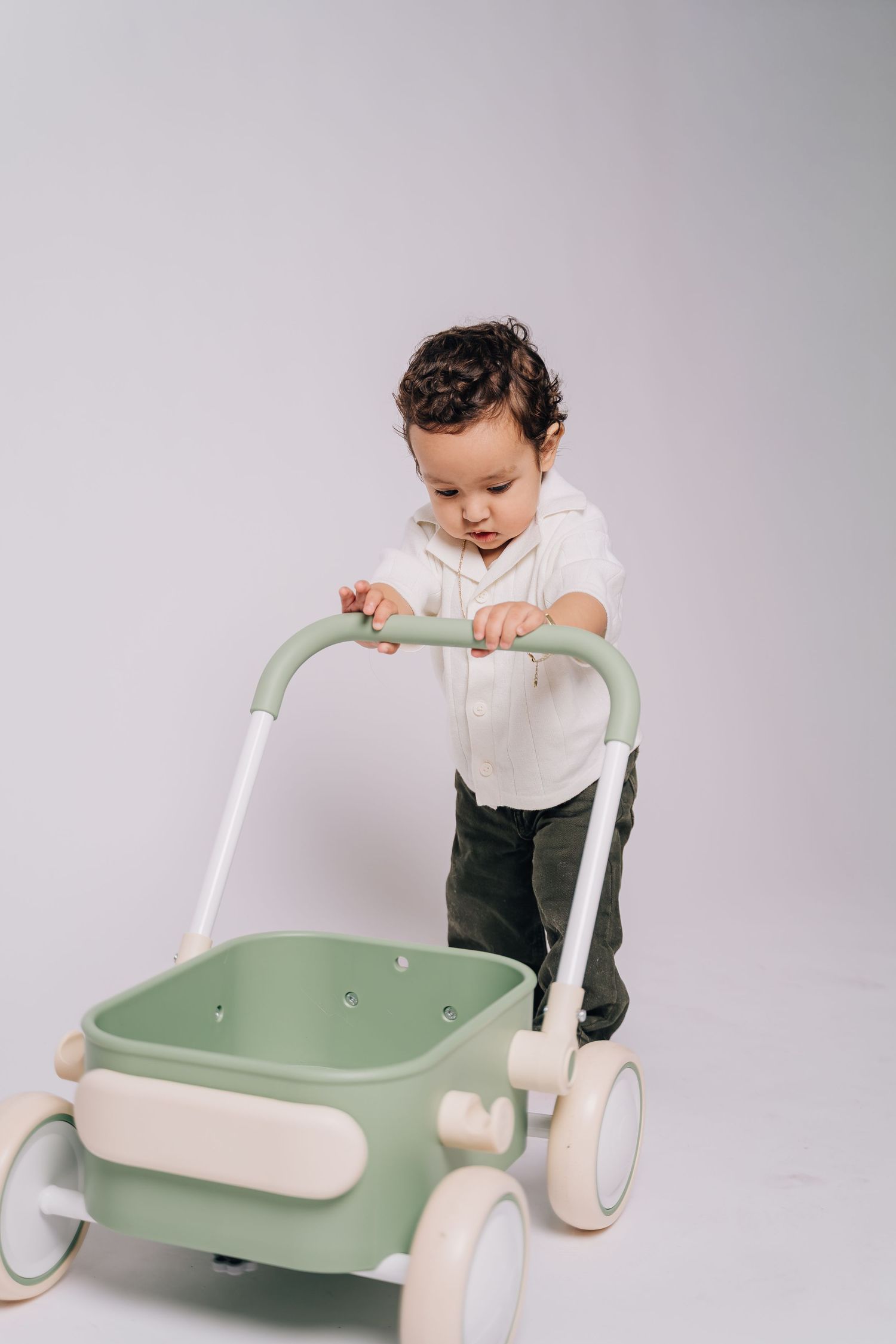Toddler in white hoodie and green pants pushing a mint green baby walker toy on a white background.