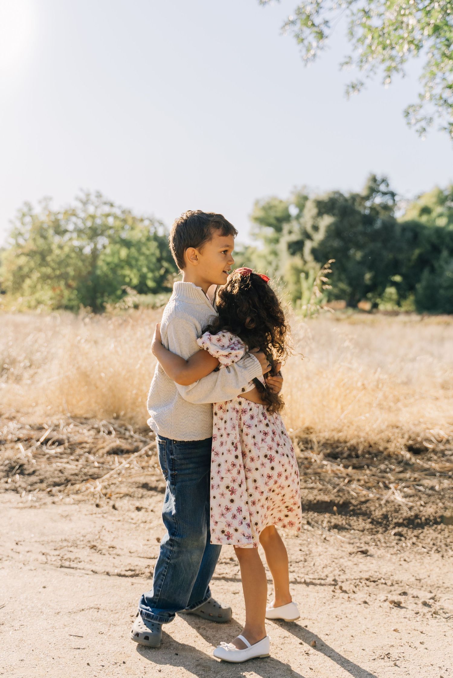 Two toddlers in casual clothing share a sweet moment together in a sunny outdoor setting with dry grass and trees.