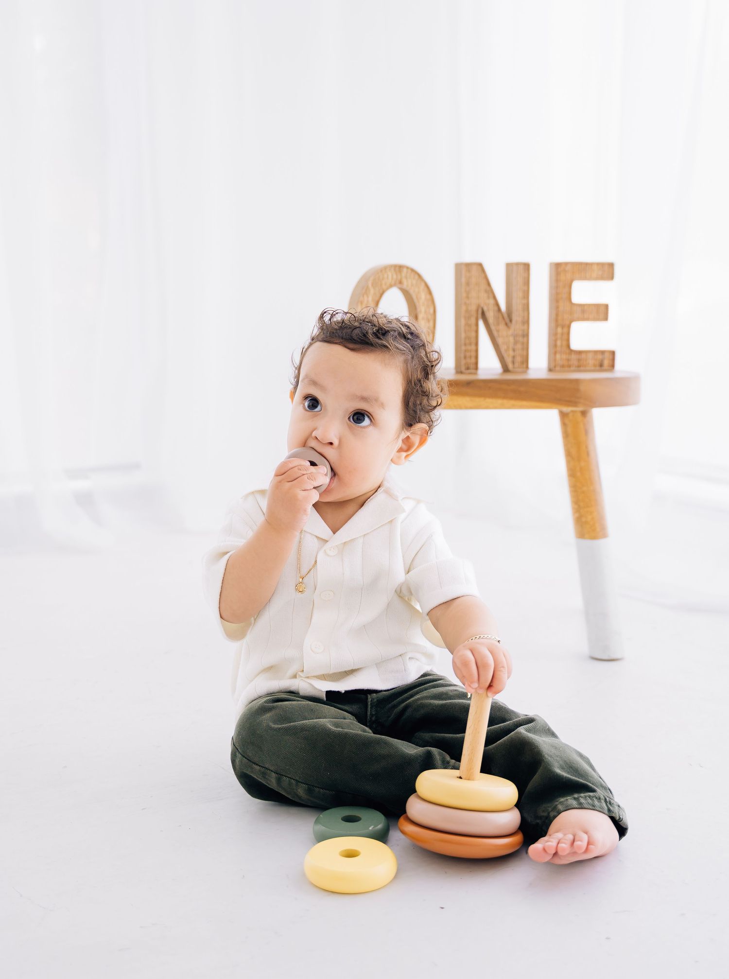 A baby boy sits on a white floor playing with a stacking toy, with a wooden ONE sign behind him.