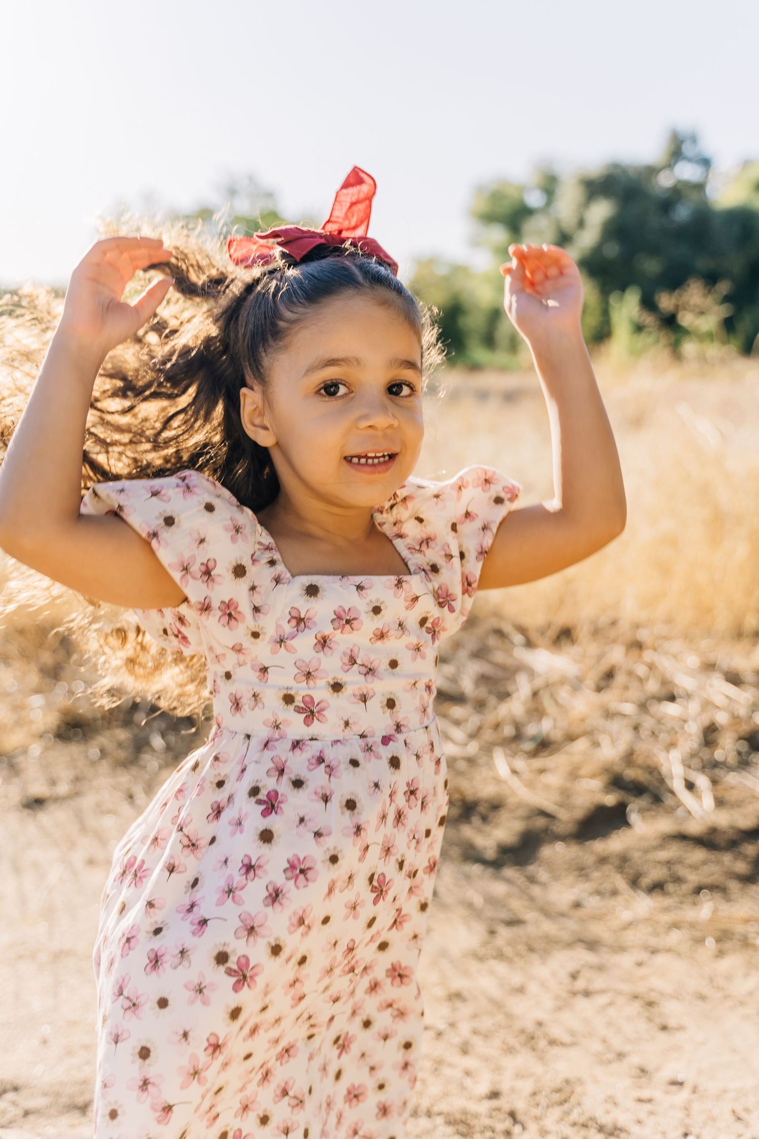 A child in a floral dress dances joyfully in a sun-drenched field with arms raised in celebration.