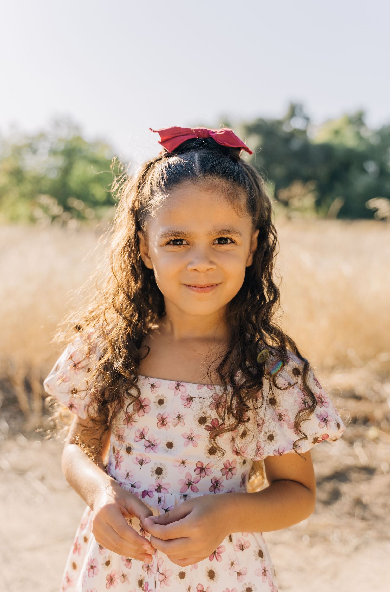 A young child in a floral dress poses in a sunlit field with natural golden grass in the background.