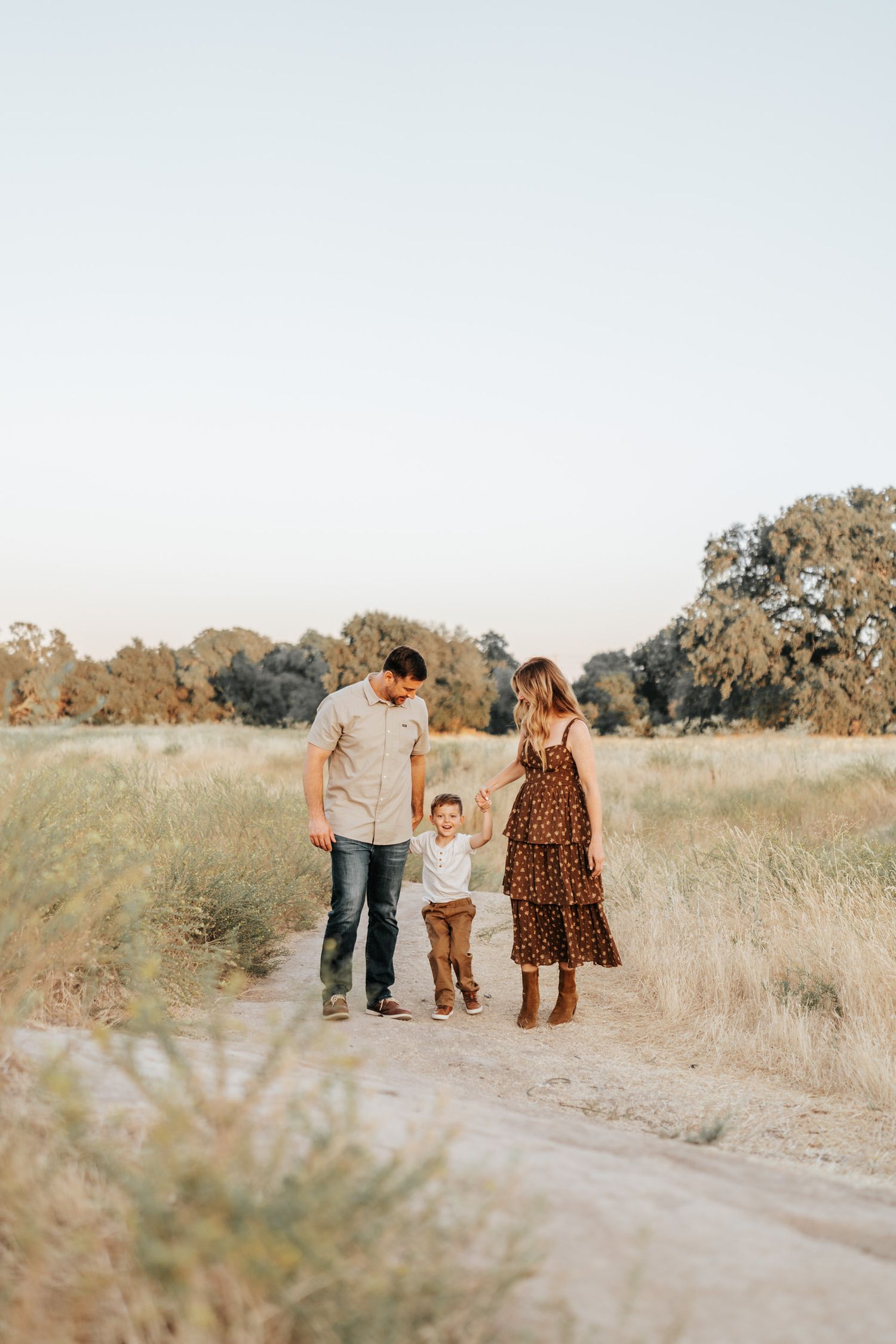 A family holds hands while walking together on a dirt path through a golden meadow.