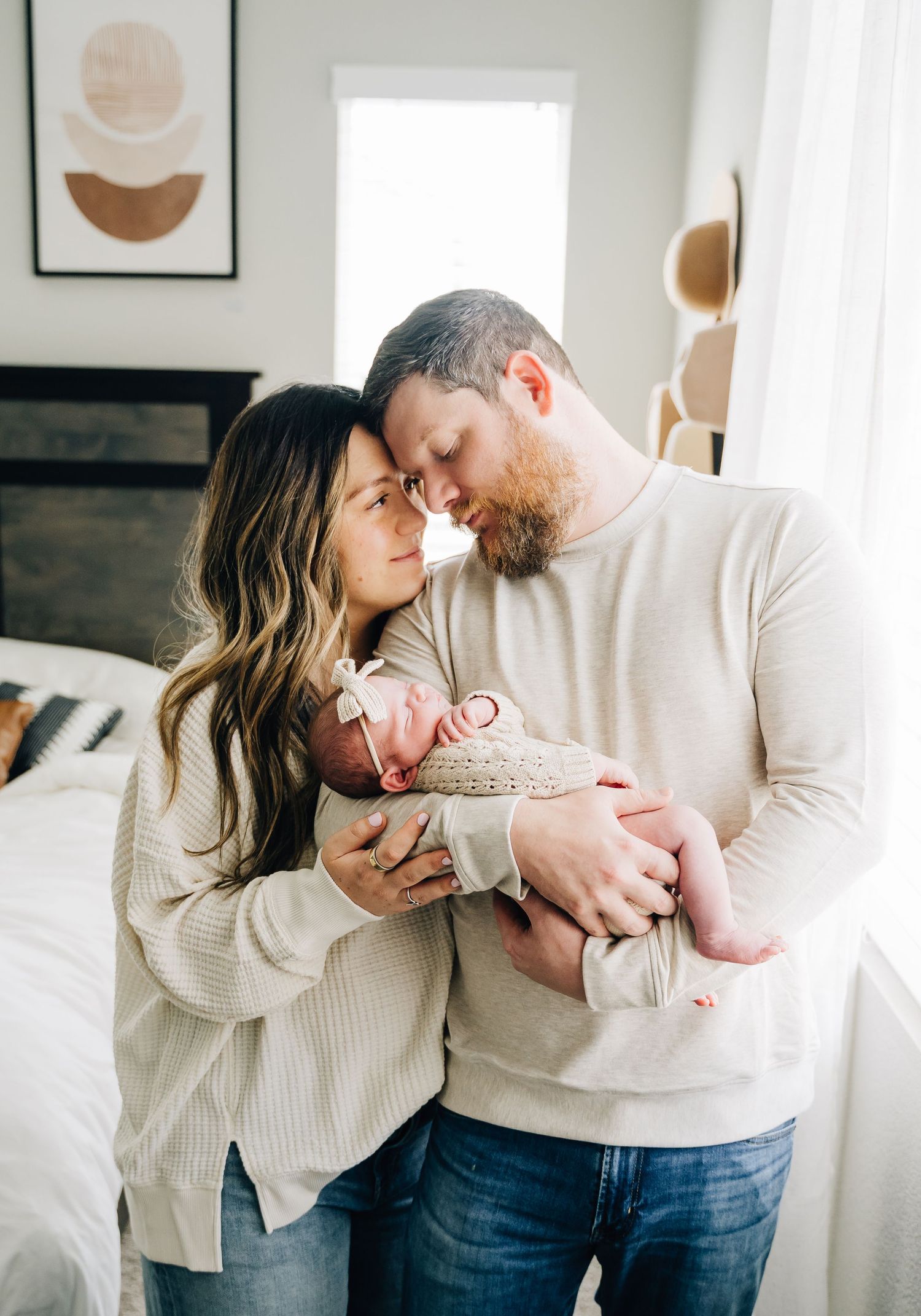 A couple in matching cream sweaters tenderly touch foreheads while cradling their sleeping newborn baby at home.