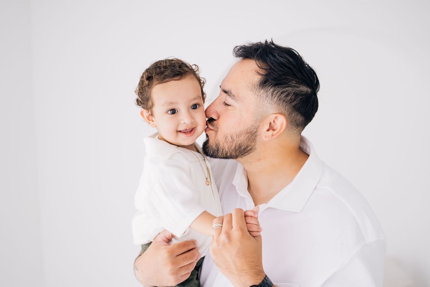 A bearded father with tattoos kisses his smiling curly-haired toddler in a bright white studio setting.
