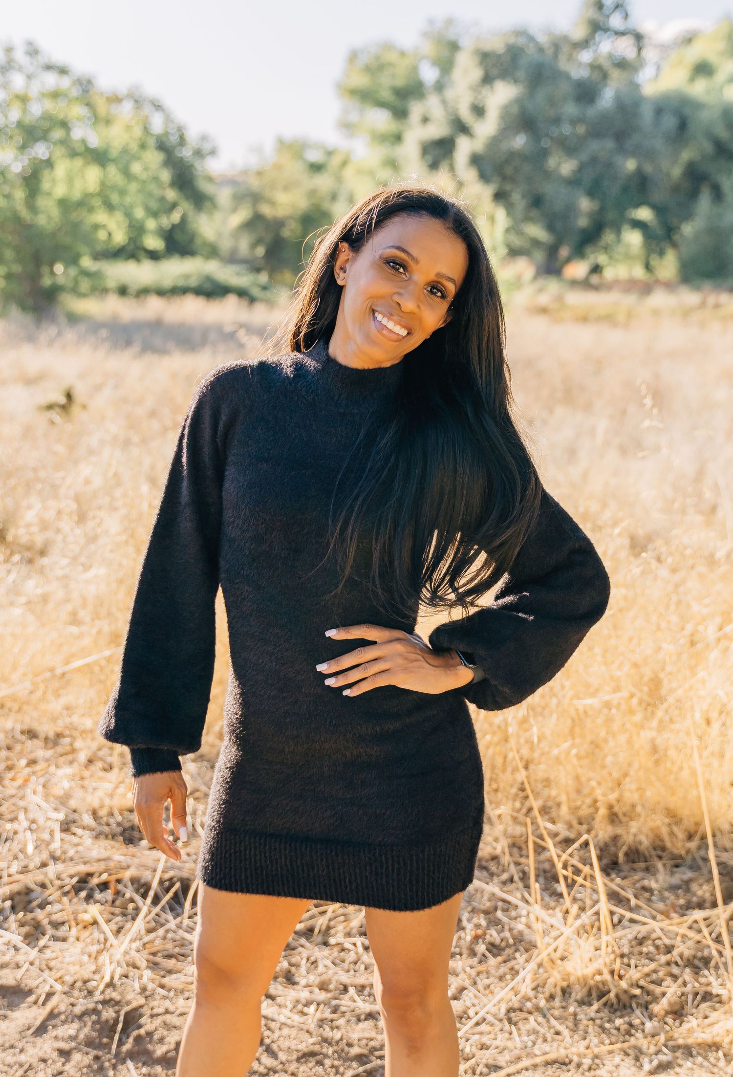 A person in a black sweater poses playfully in a golden field during sunset.