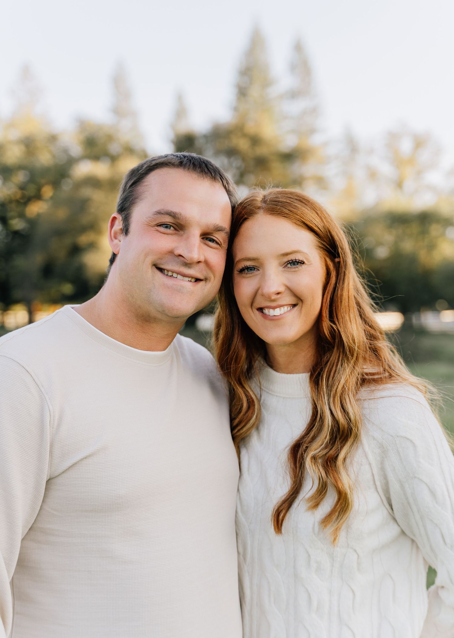 A couple wearing matching white sweaters poses together outdoors during golden hour.