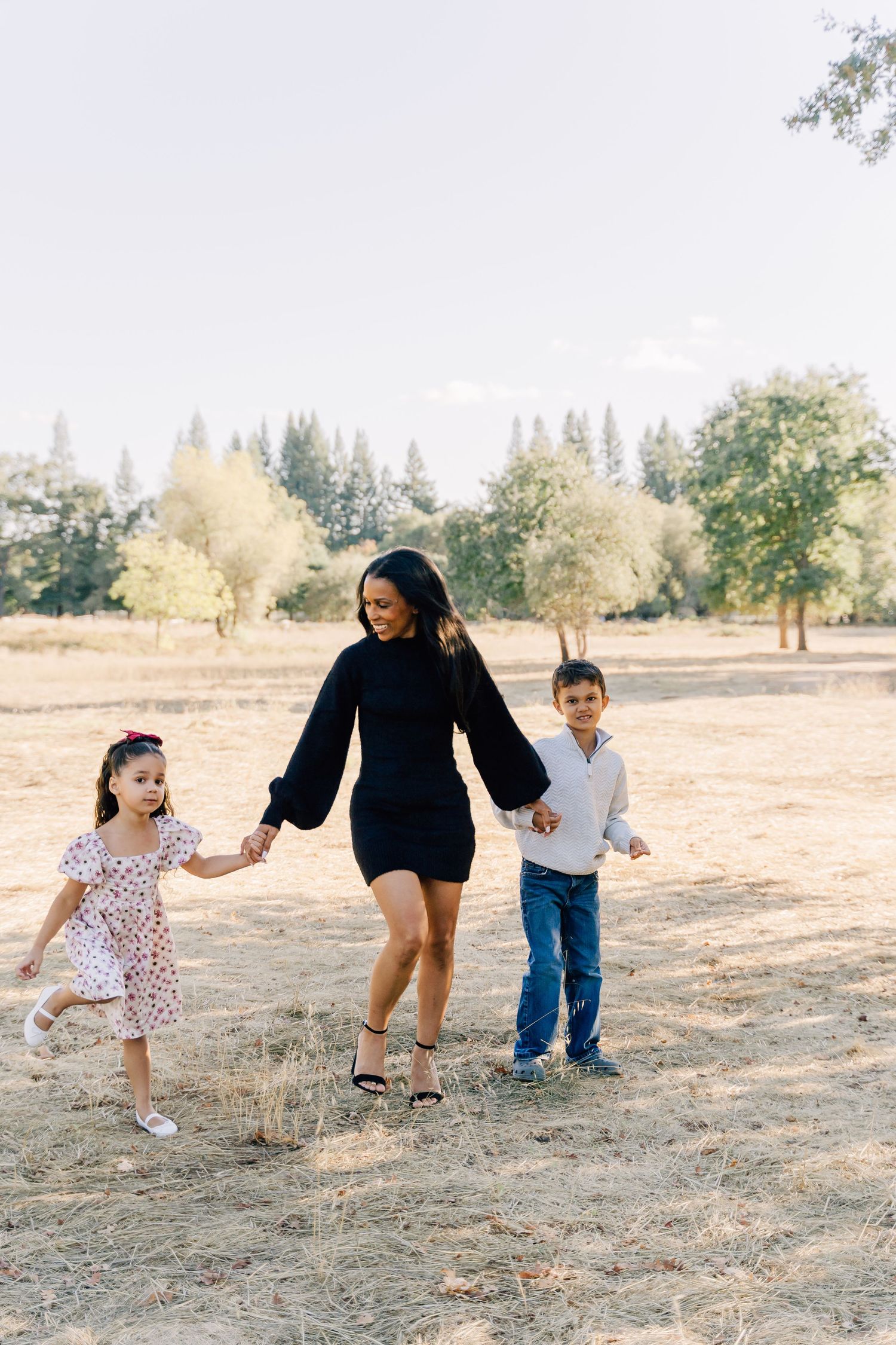 A person in a black dress walks hand in hand with two children through a sunny park with trees in the background.
