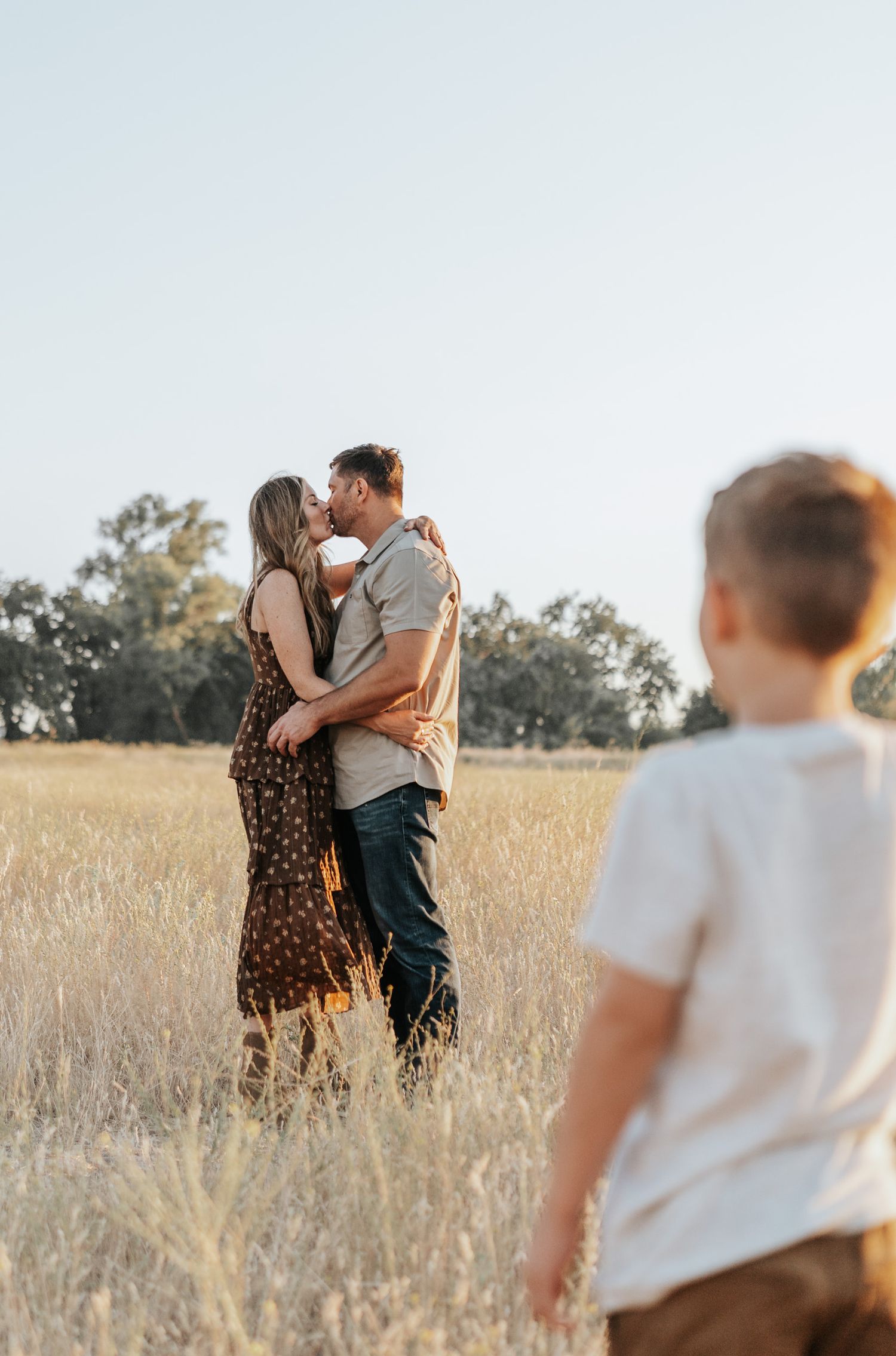 A couple embraces in a golden wheat field at sunset while a child watches from the side.