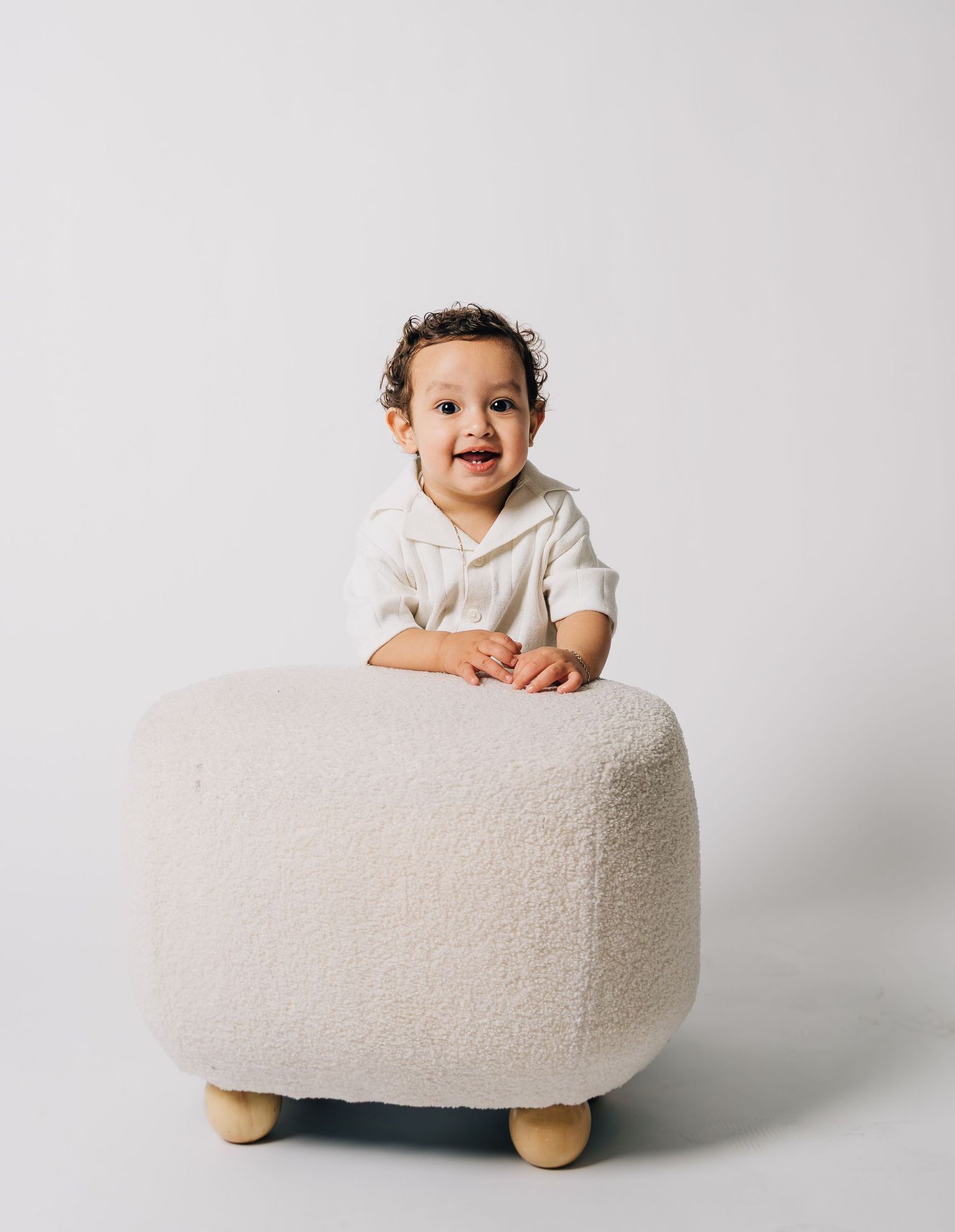 Happy baby in cream romper standing at a fluffy white ottoman in a bright studio setting.