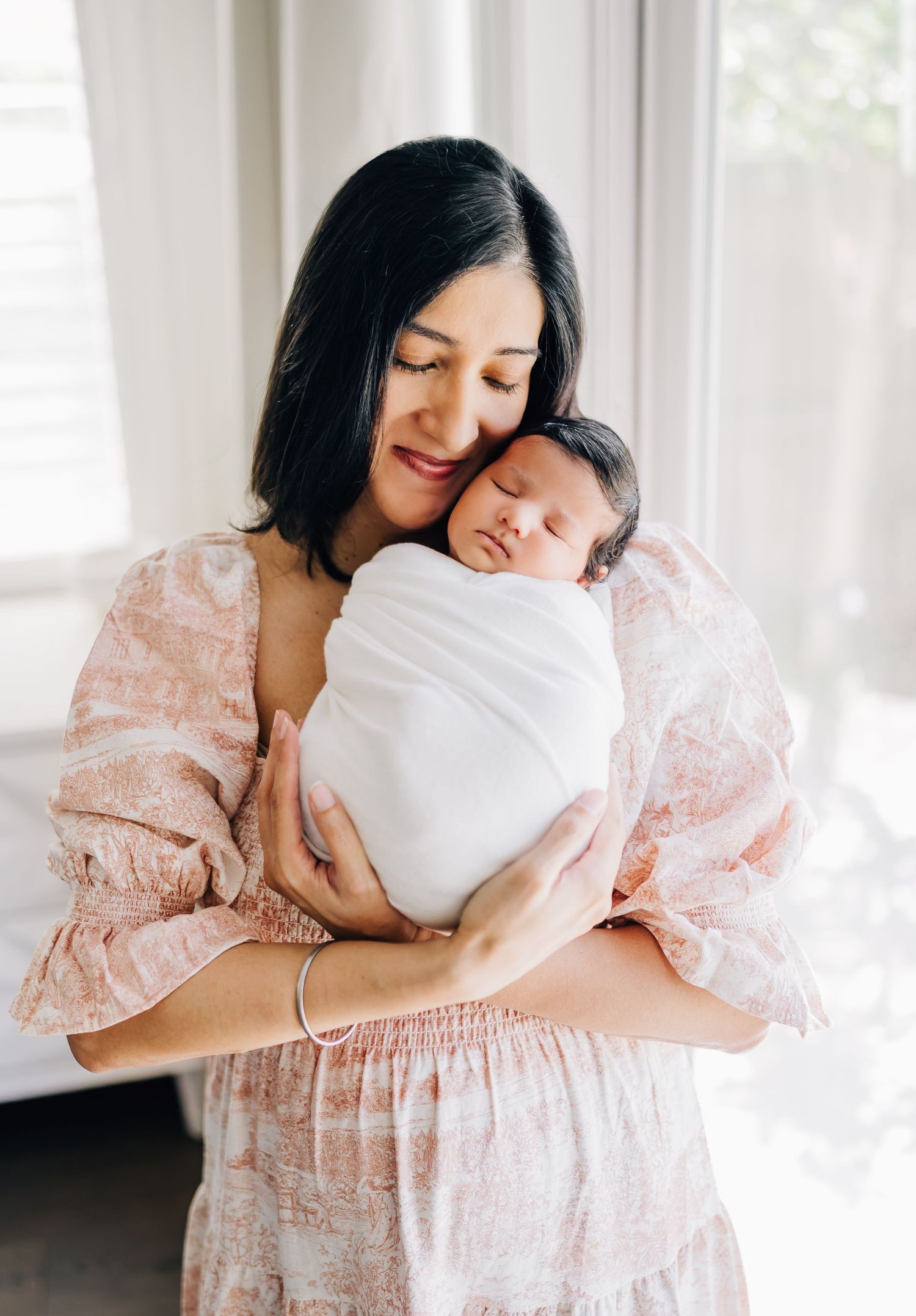 A mother in a floral dress tenderly holds her swaddled newborn baby close to her cheek near a bright window.