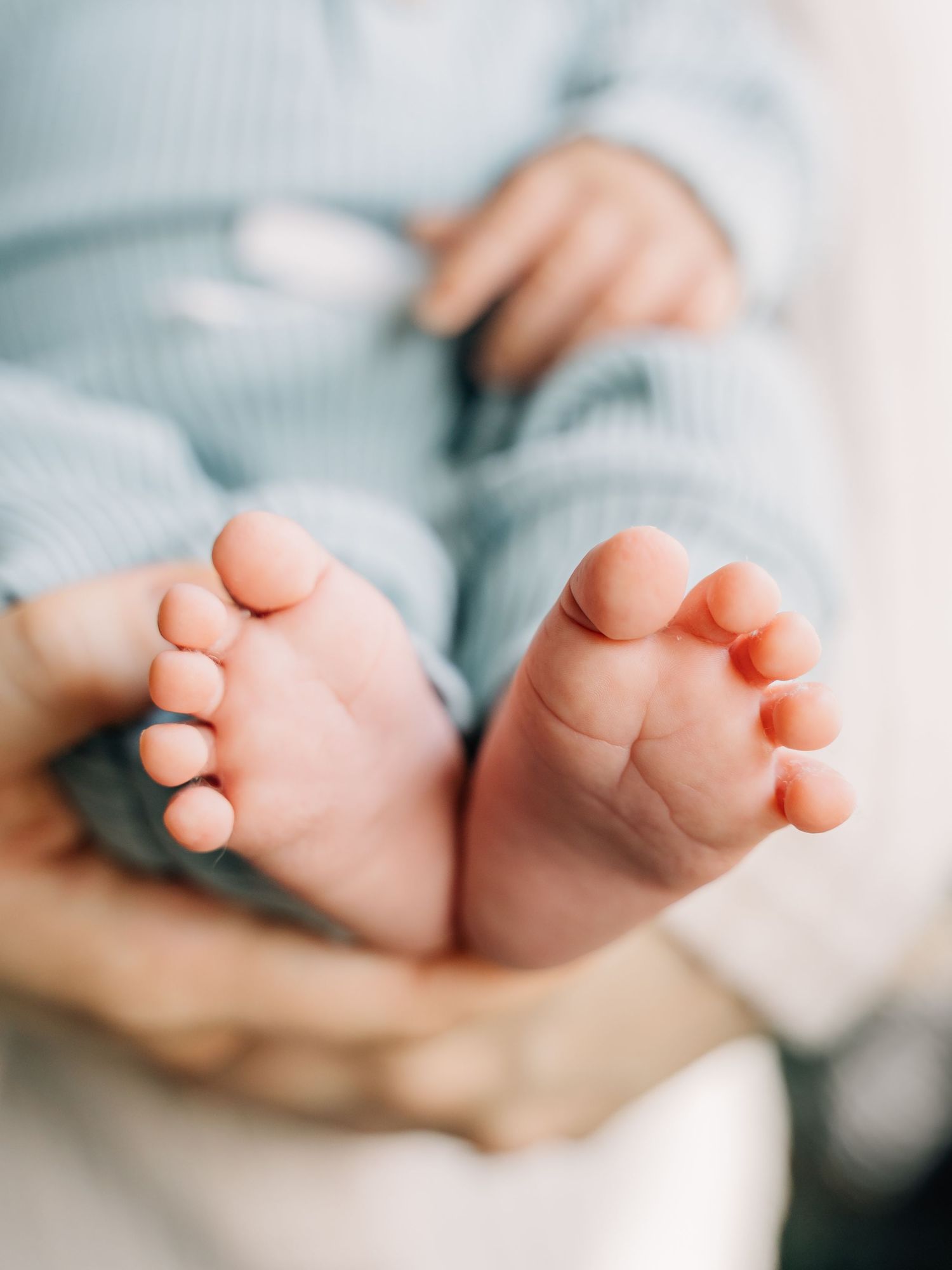 Close-up of tiny newborn baby feet held gently in adult hands, baby wearing a soft blue onesie.