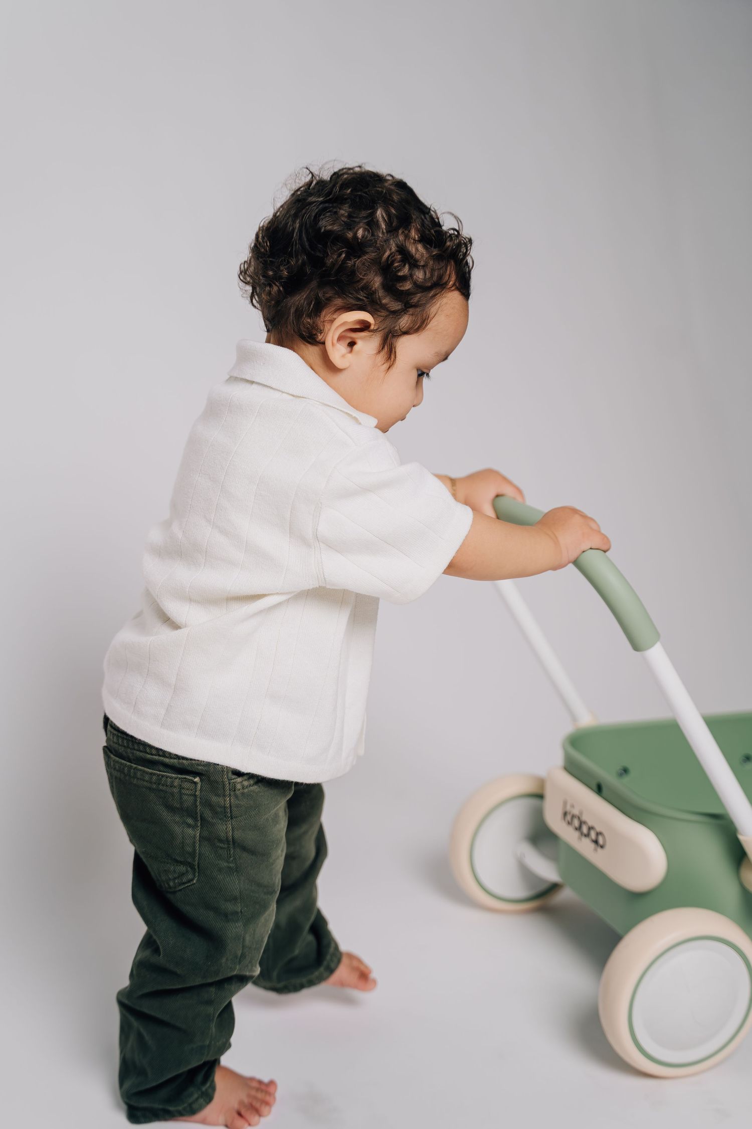 Toddler in white shirt and green pants pushing a wooden baby walker toy on white background.
