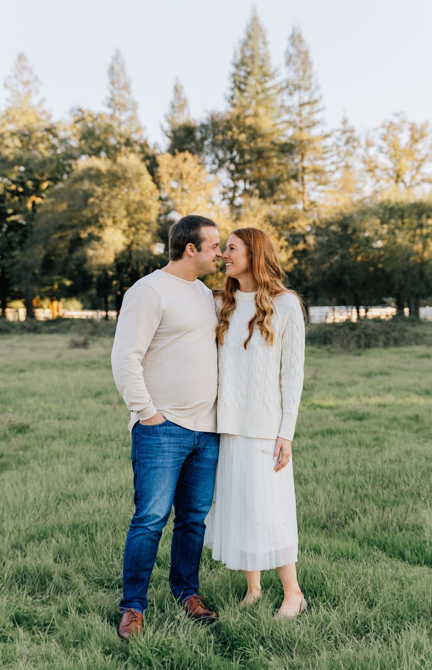 A couple in casual attire sharing romantic moments in a grassy field with evergreen trees in the background.