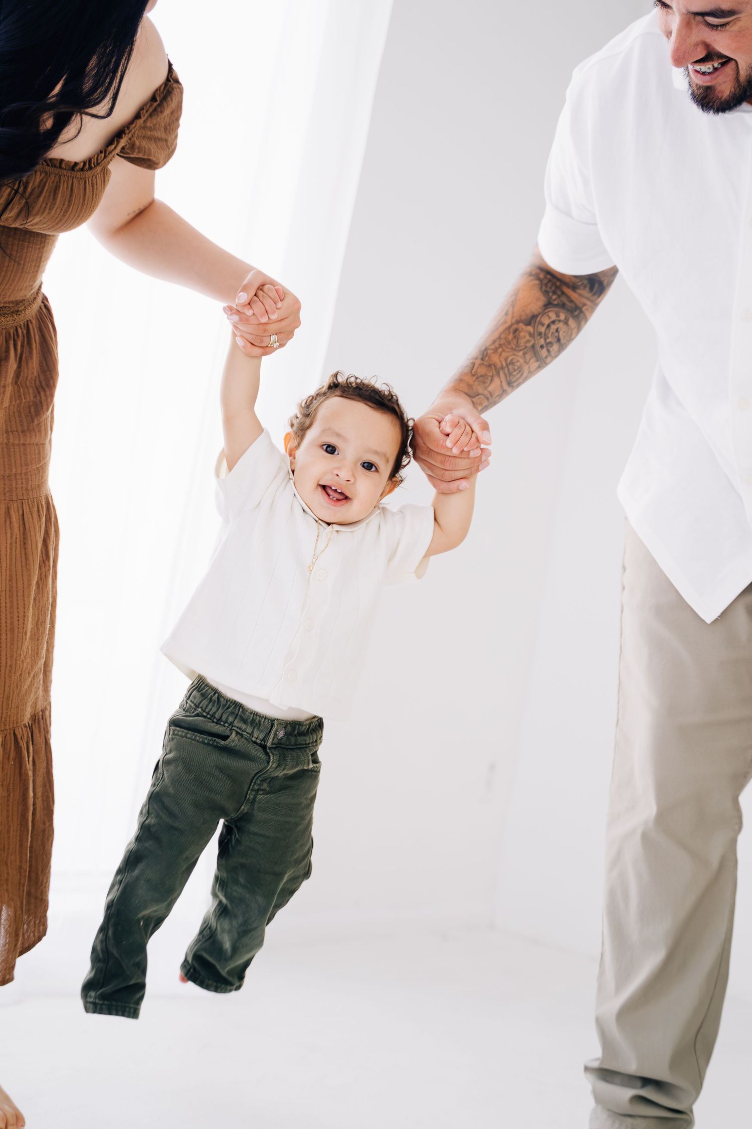 Parents holding hands of smiling baby taking first steps in white studio, family milestone moment.