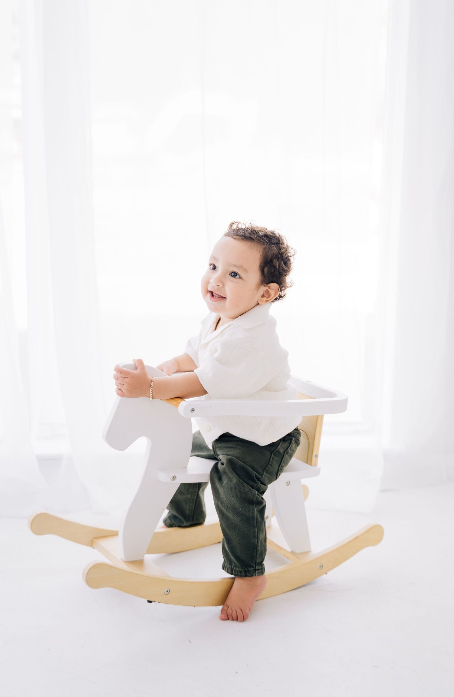 Smiling toddler in white shirt and green pants sitting on a white wooden rocking horse toy.