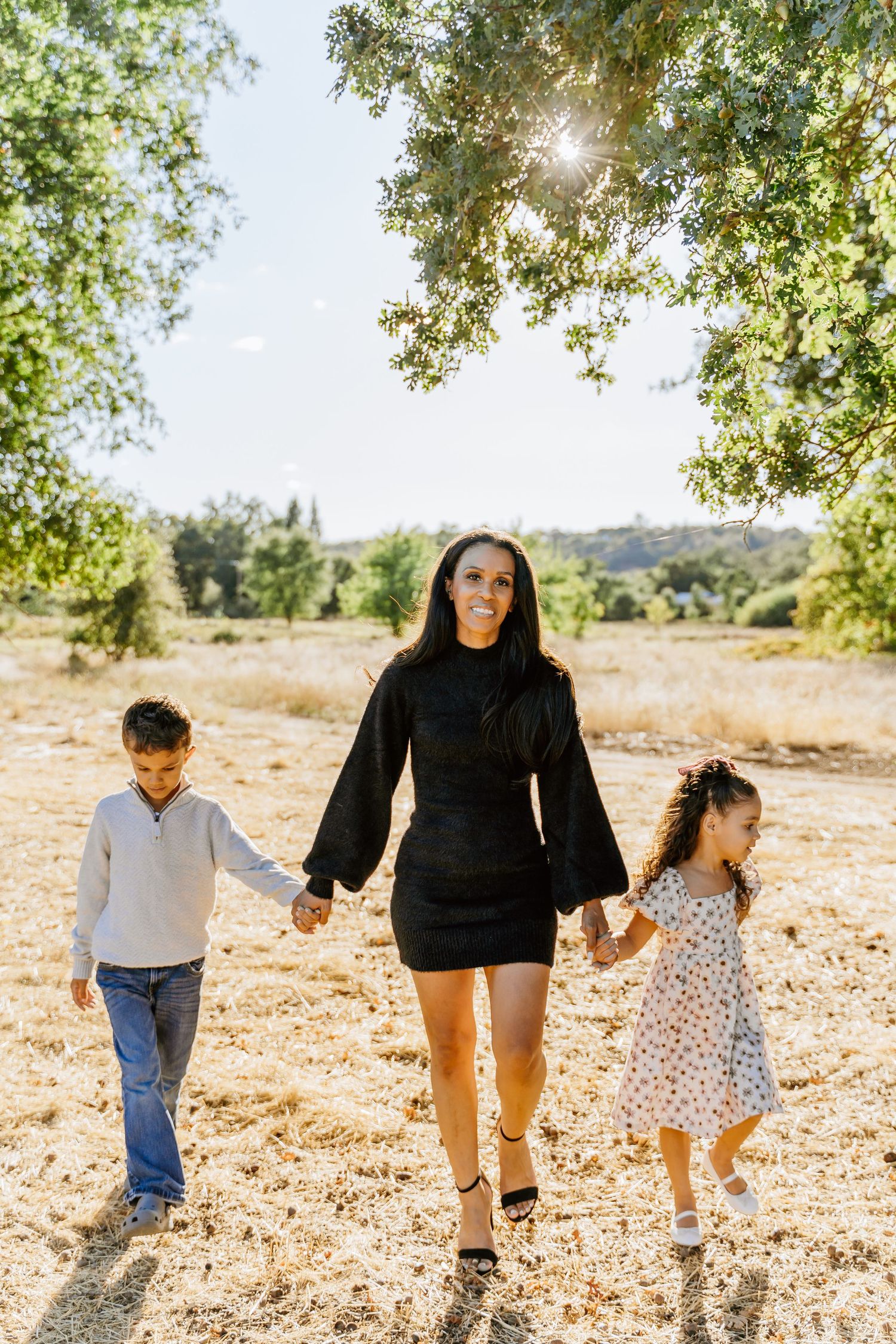 Two children and an adult walk together on a dusty path under trees on a sunny day.