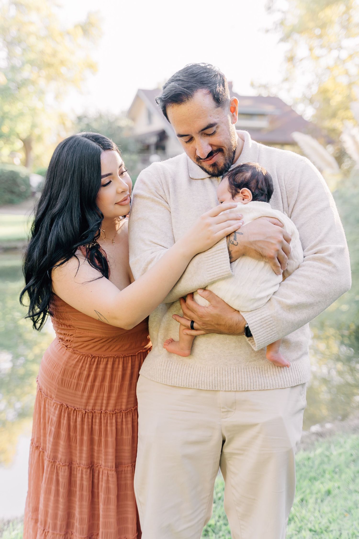 A couple in warm tones smiles lovingly at their newborn baby outdoors near a pond.