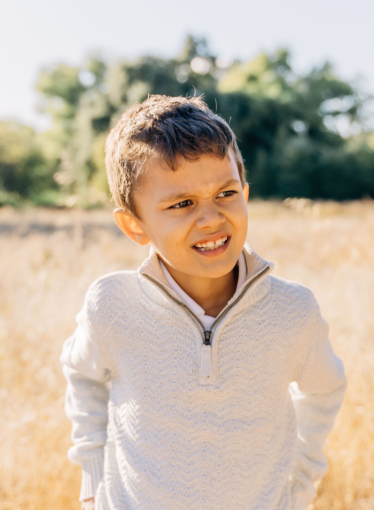 Child wearing a cream colored sweater smiles warmly in a sun-drenched field of tall grass.