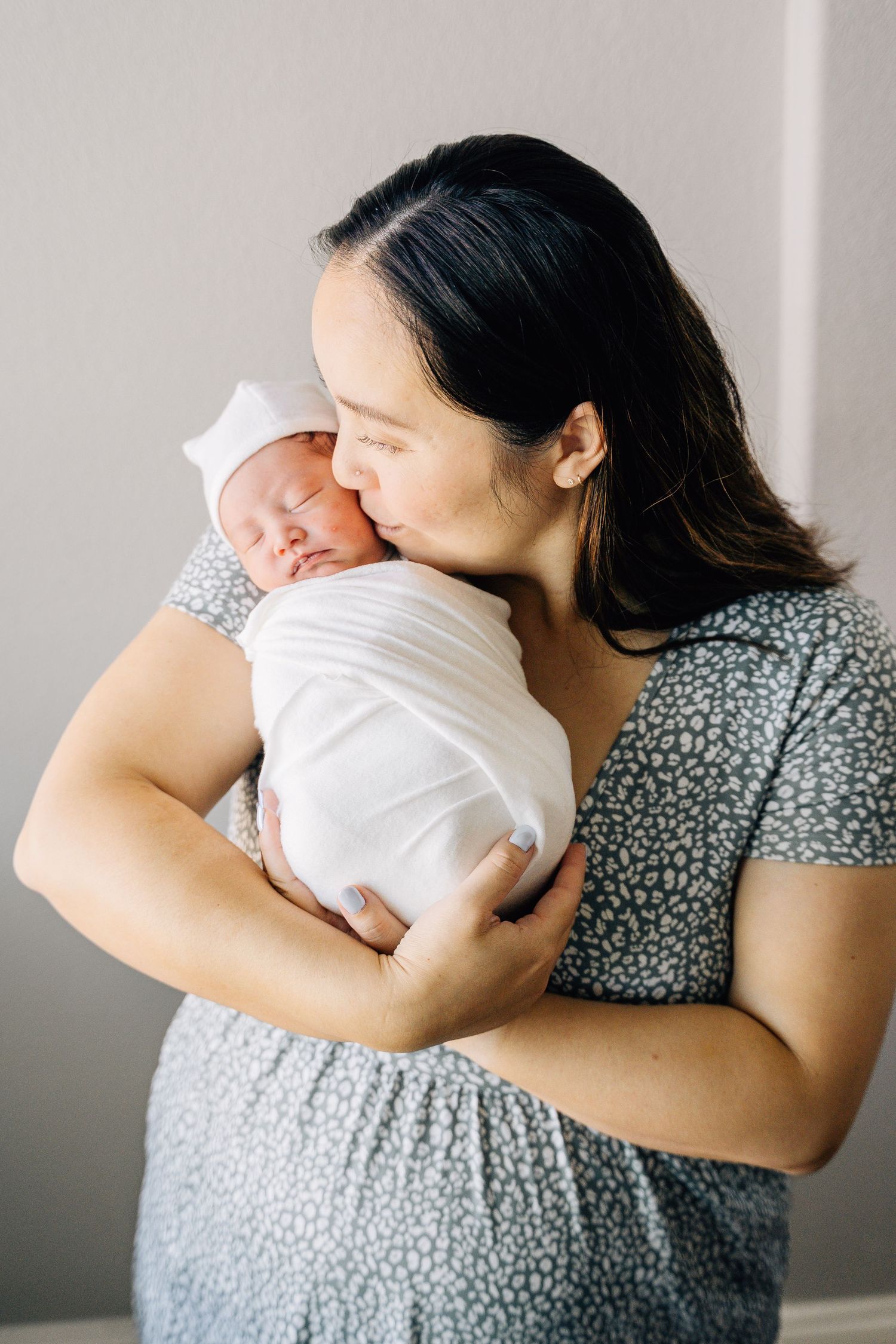 A mother tenderly kisses her swaddled newborn baby wrapped in white, wearing a floral dress.