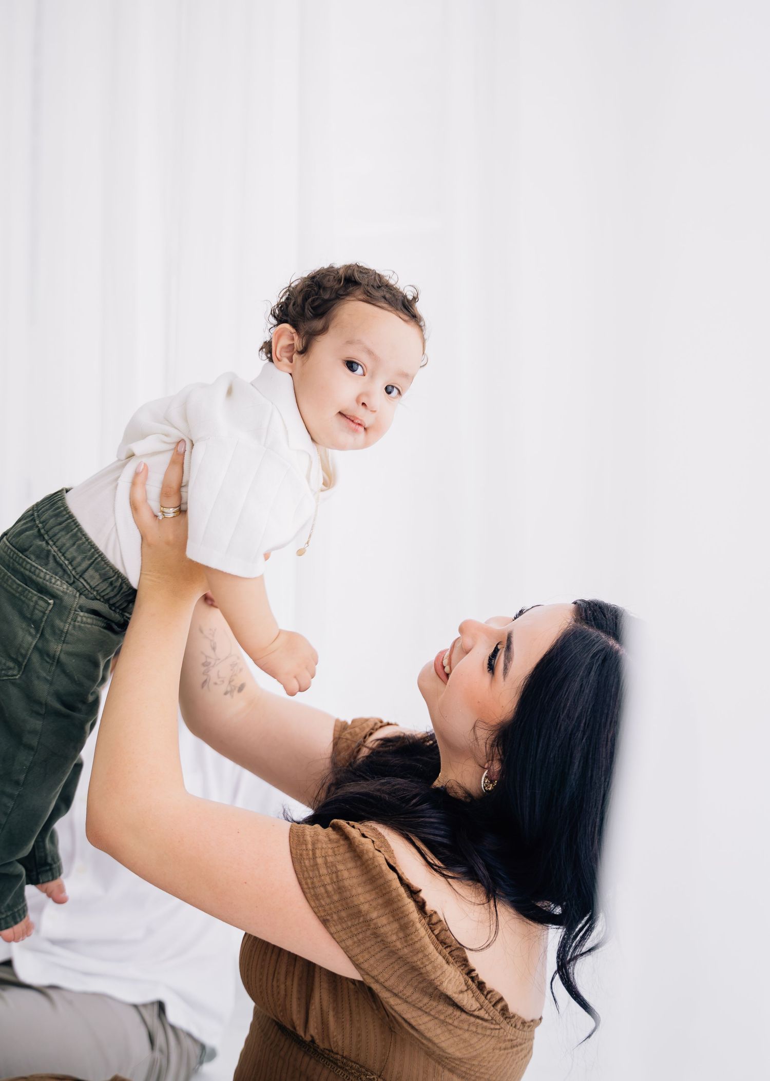 A joyful mother lifts her smiling baby boy in a bright white studio, both sharing a loving gaze.