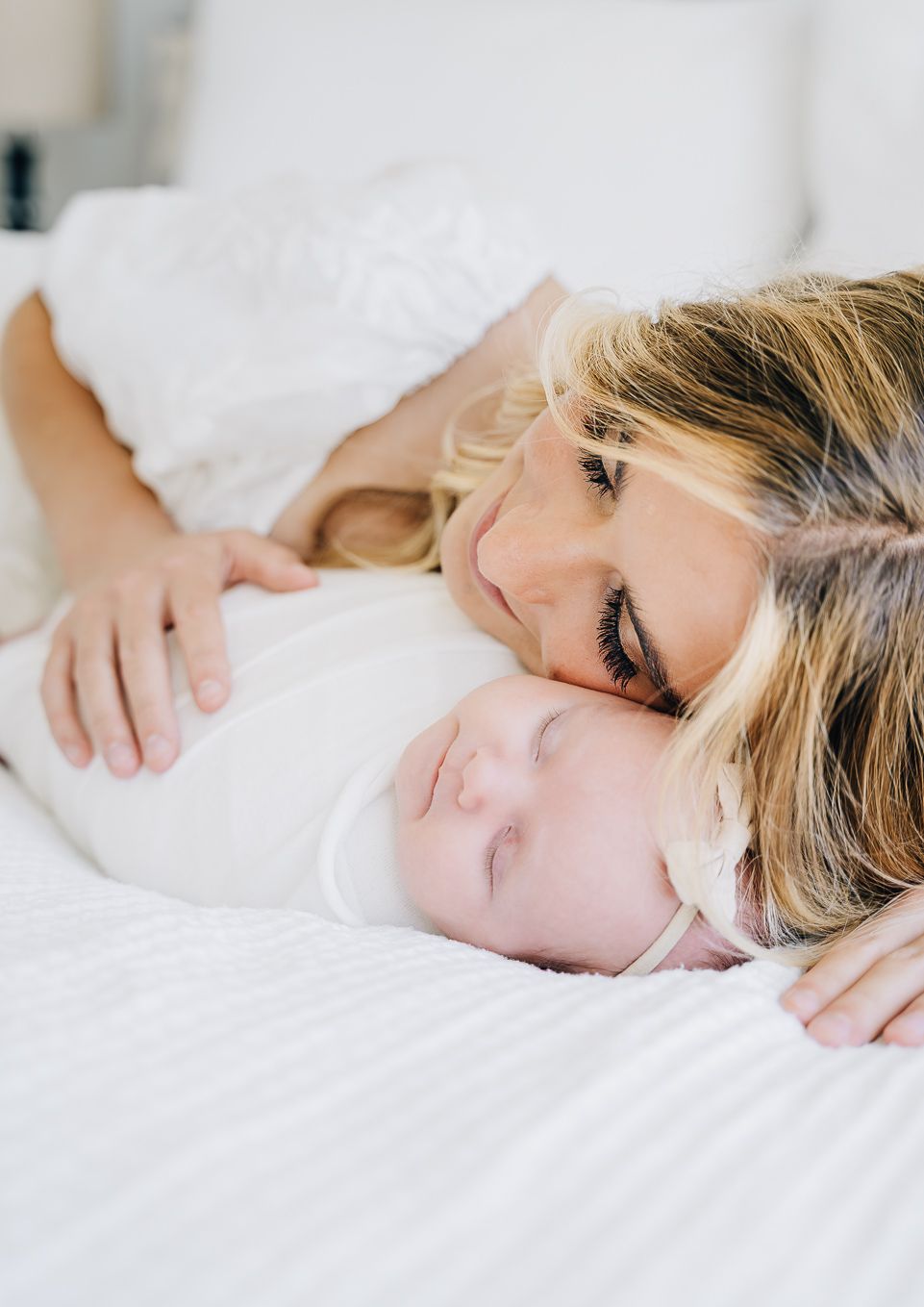 A mother tenderly rests her cheek against her sleeping newborn baby on a white bed.