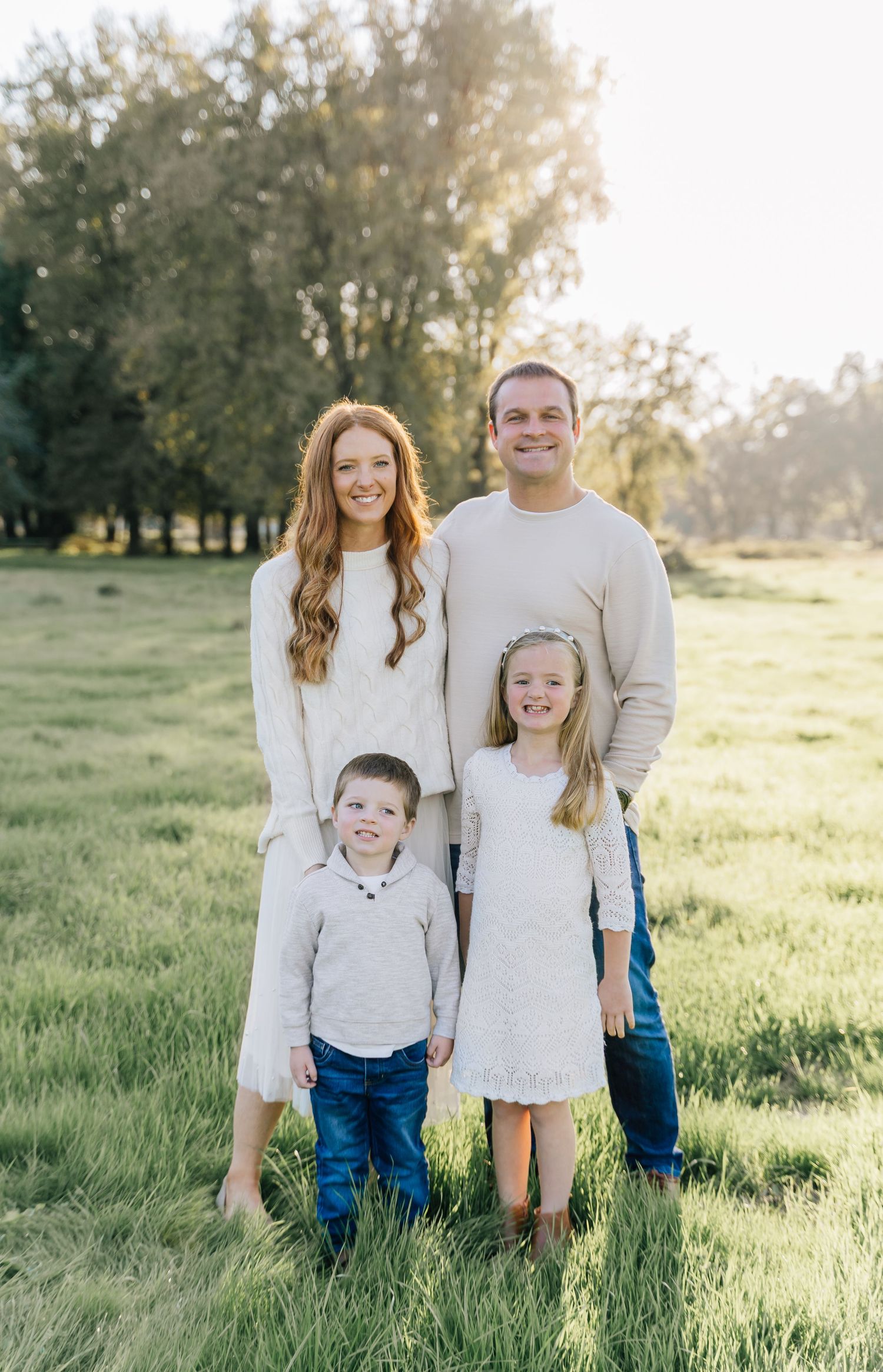 A family wearing white and blue outfits poses together in a sunlit grassy field during golden hour.