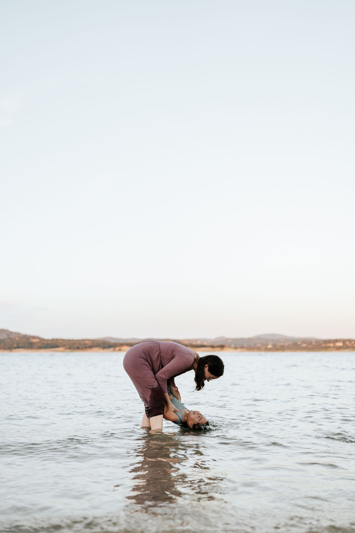 Person bends down searching in shallow ocean water near a rocky beach shore.