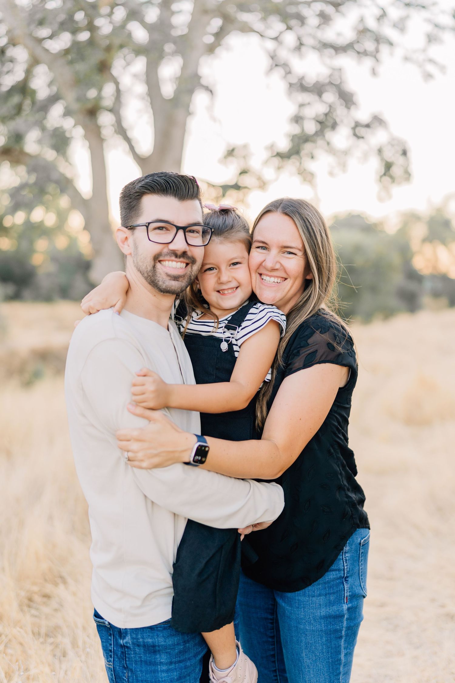 Happy family of three posing outdoors in a golden field, parents hugging their young daughter, smiling warmly.