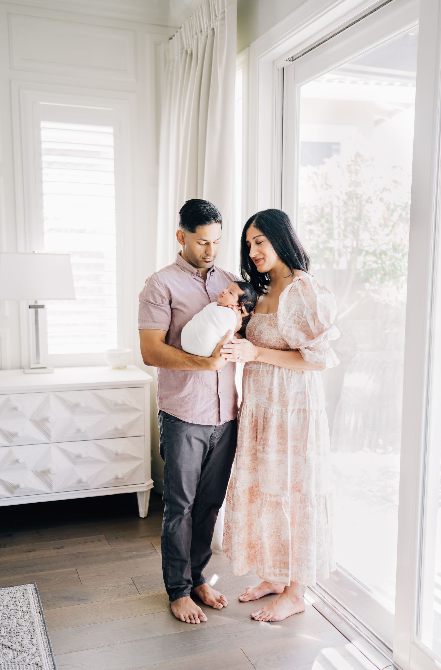 A couple in a bright nursery gazes lovingly at their newborn baby wrapped in white near large windows.