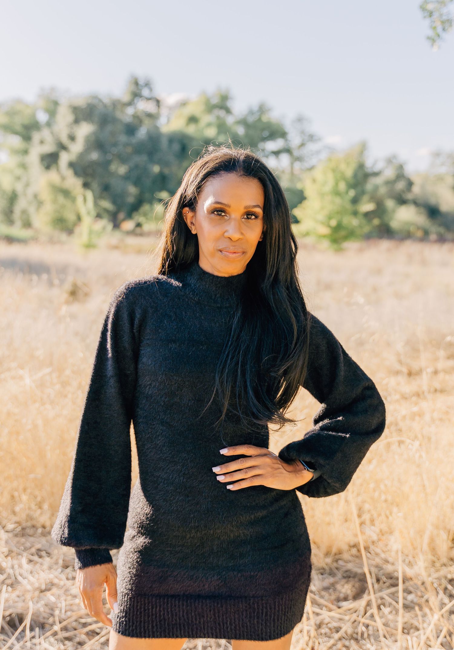A woman in a black sweater poses confidently against a golden field backdrop during a sunny outdoor photoshoot.