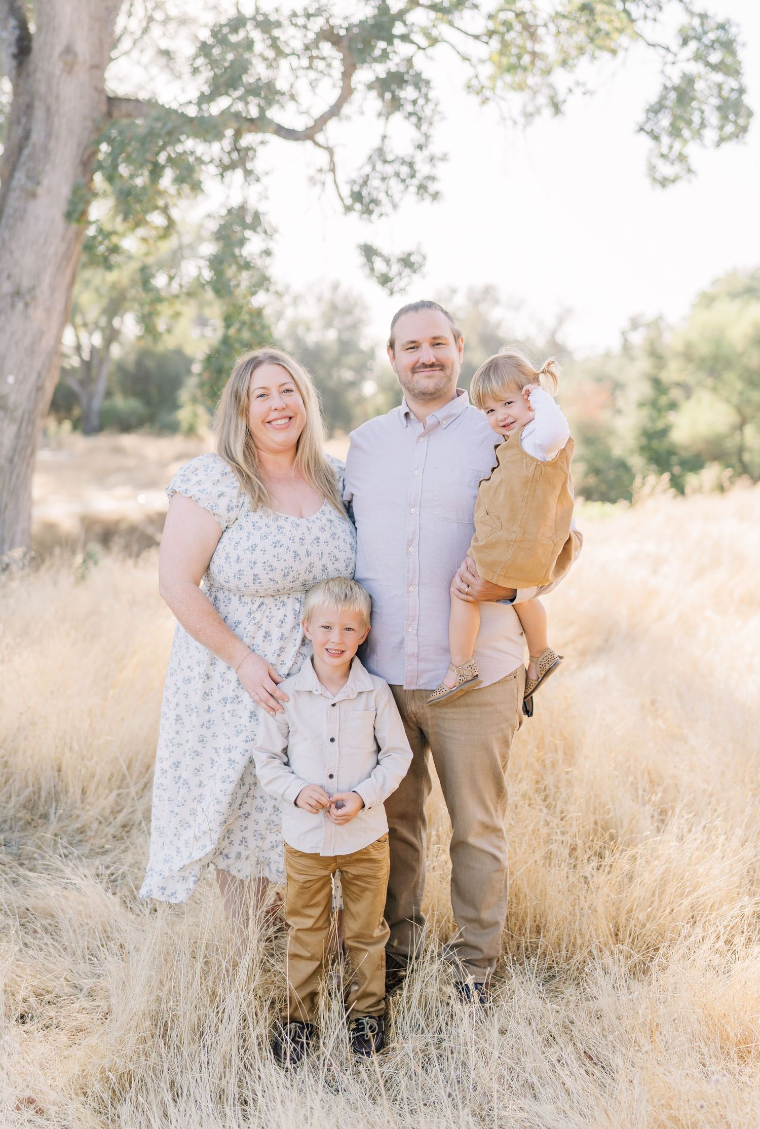 Family of four posing outdoors in golden field with oak trees, parents smiling with two young boys on sunny day.