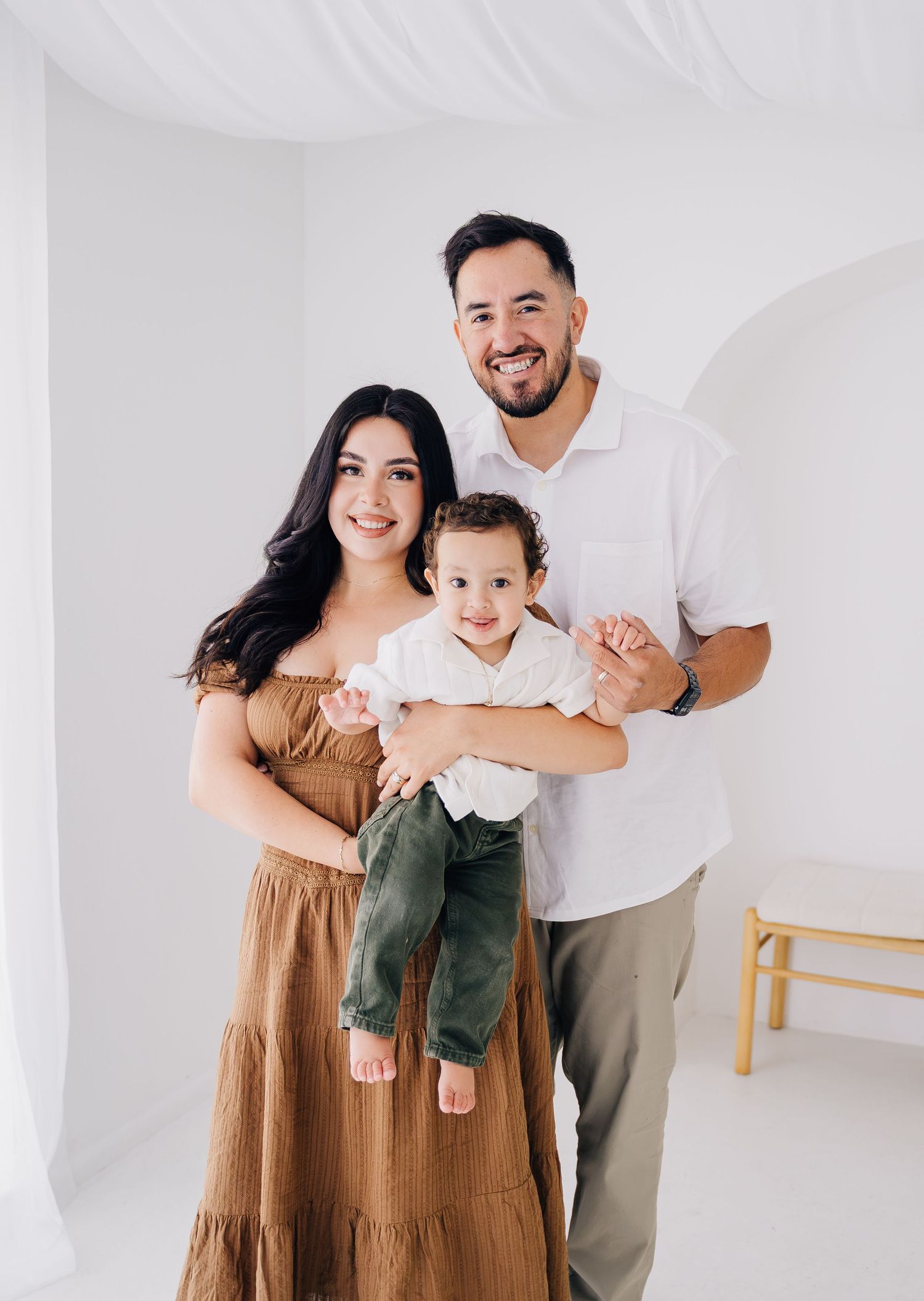 A smiling family of three poses in a bright white studio, parents holding their toddler son.
