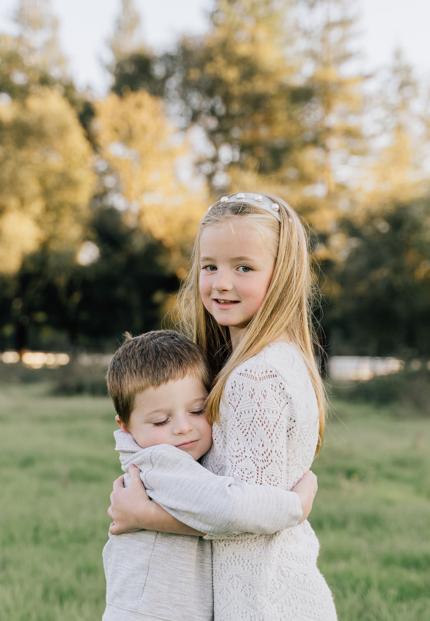 Two children in white outfits share a sweet hug outdoors on a sunny autumn day in a grassy field.