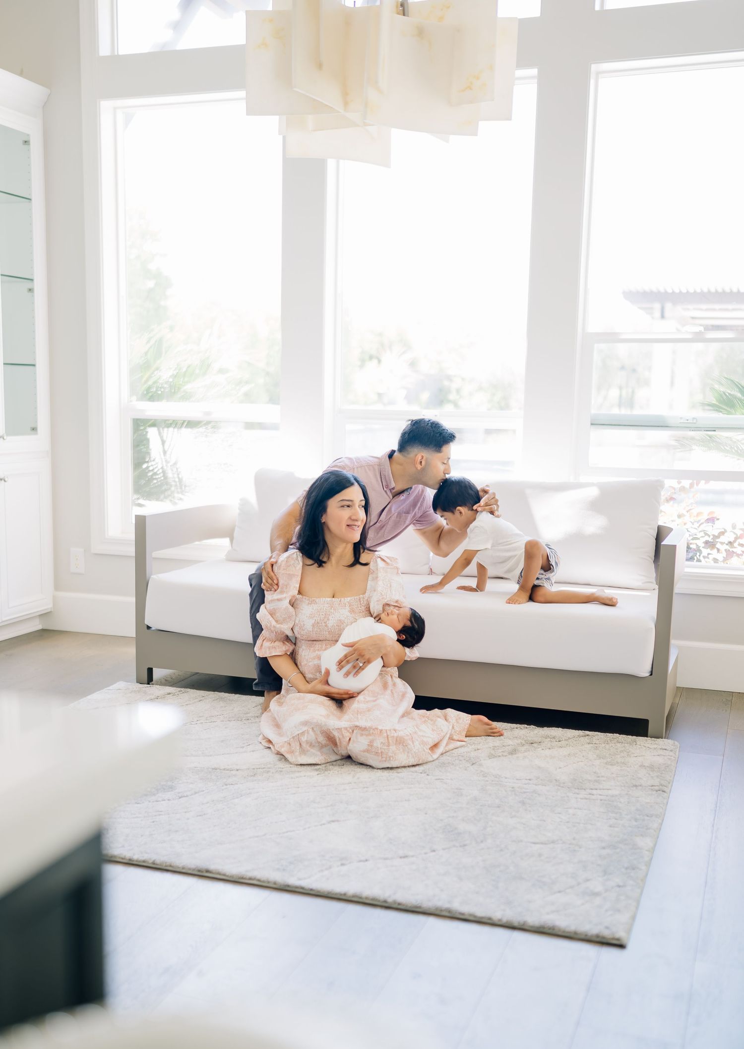 A family poses in a bright living room, mom sitting on a rug holding a newborn while dad and child relax on a white sofa.